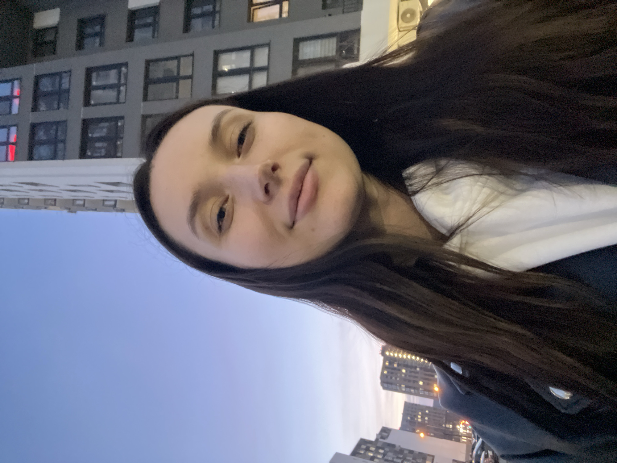 A woman is taking a selfie outdoors at dusk in Tyumen, Russia. She has long, dark hair, light skin, and is wearing a white hooded sweatshirt under a dark jacket. Her face is the central focus of the image. Behind her, a gray building with multiple windows is visible on one side, while on the other side, the sky is a gradient of blue to orange, with the silhouettes of several buildings visible on the horizon. The scene suggests an urban environment with residential or commercial buildings.