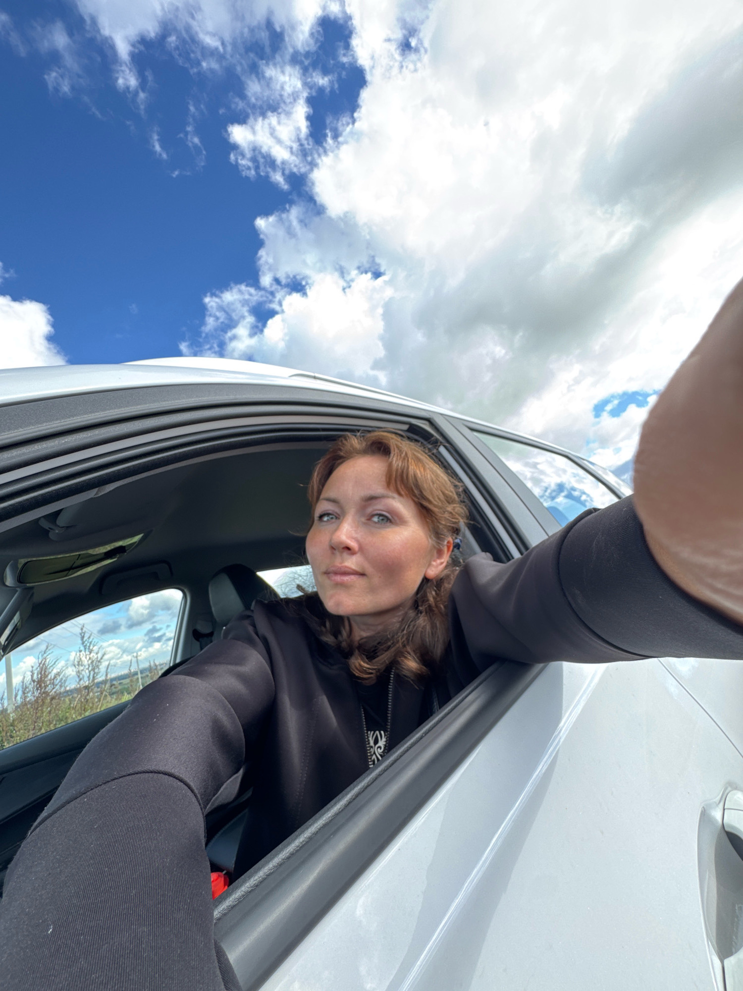 A woman with auburn hair and blue eyes is taking a selfie from inside a car. She is wearing a black hoodie and has her left arm resting on the open car door, with her hand blurred in the foreground. The car is light-colored. Outside the car, a bright blue sky with white clouds is visible, along with some dry vegetation in the background. The image appears to have been taken outdoors during the day.