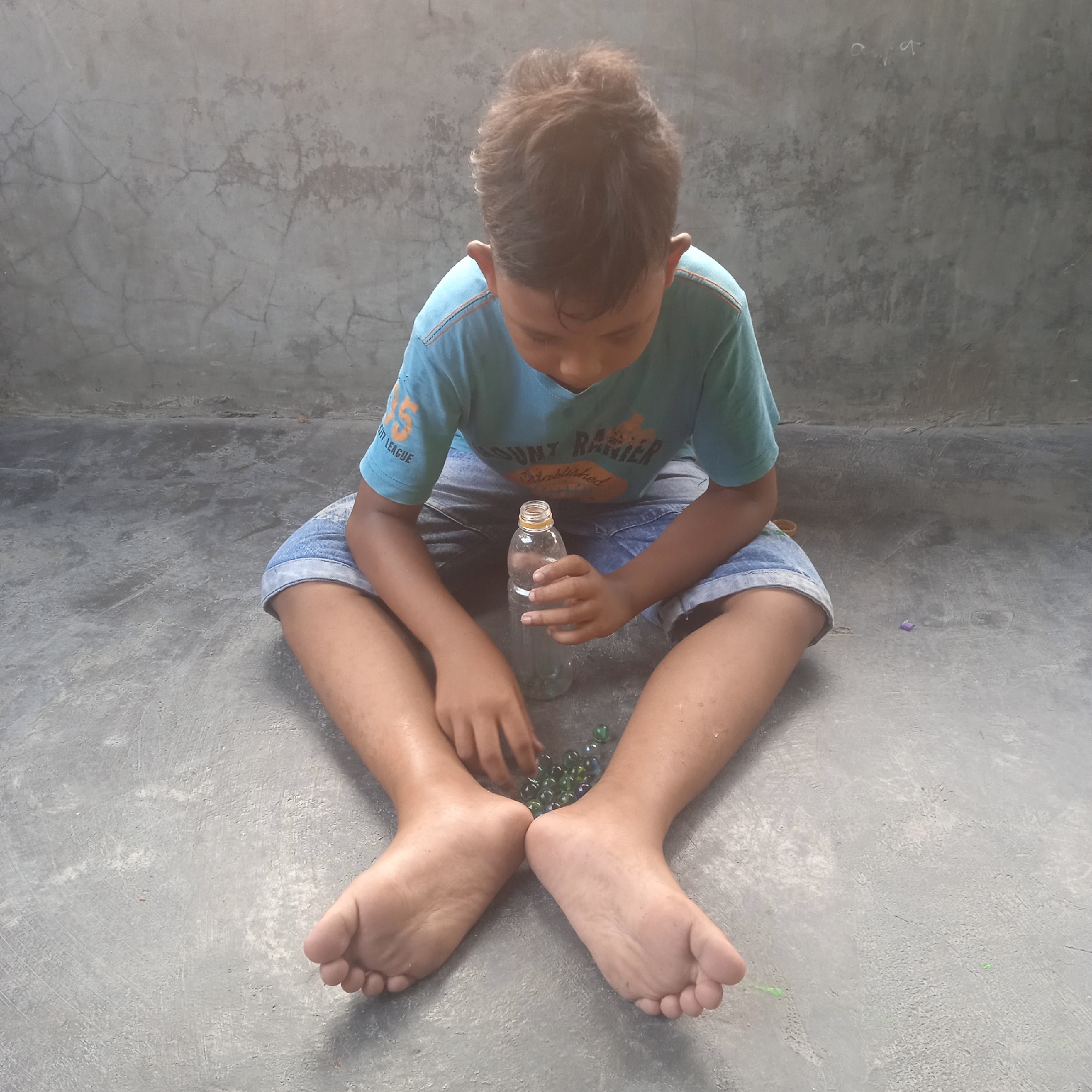 A young boy sits cross-legged on a concrete floor, facing a wall with a cracked texture. He is wearing a light blue t-shirt with orange lettering and denim shorts. In front of him, he holds a clear plastic bottle, its cap slightly ajar, and to his right, a cluster of small, colorful marbles are scattered on the ground. His hands are positioned to interact with the marbles. The lighting suggests it is daytime, possibly late afternoon given the soft, diffused light. The overall mood appears to be one of quiet concentration or play. Some of the visible text on the boy's shirt reads "MOUNT RAINIER" and "ESTABLISHED". Another part of the text on his sleeve is "FLY LEAGUE". The setting seems to be an outdoor or semi-outdoor space, perhaps a yard or a veranda, in Situterate, Indonesia.