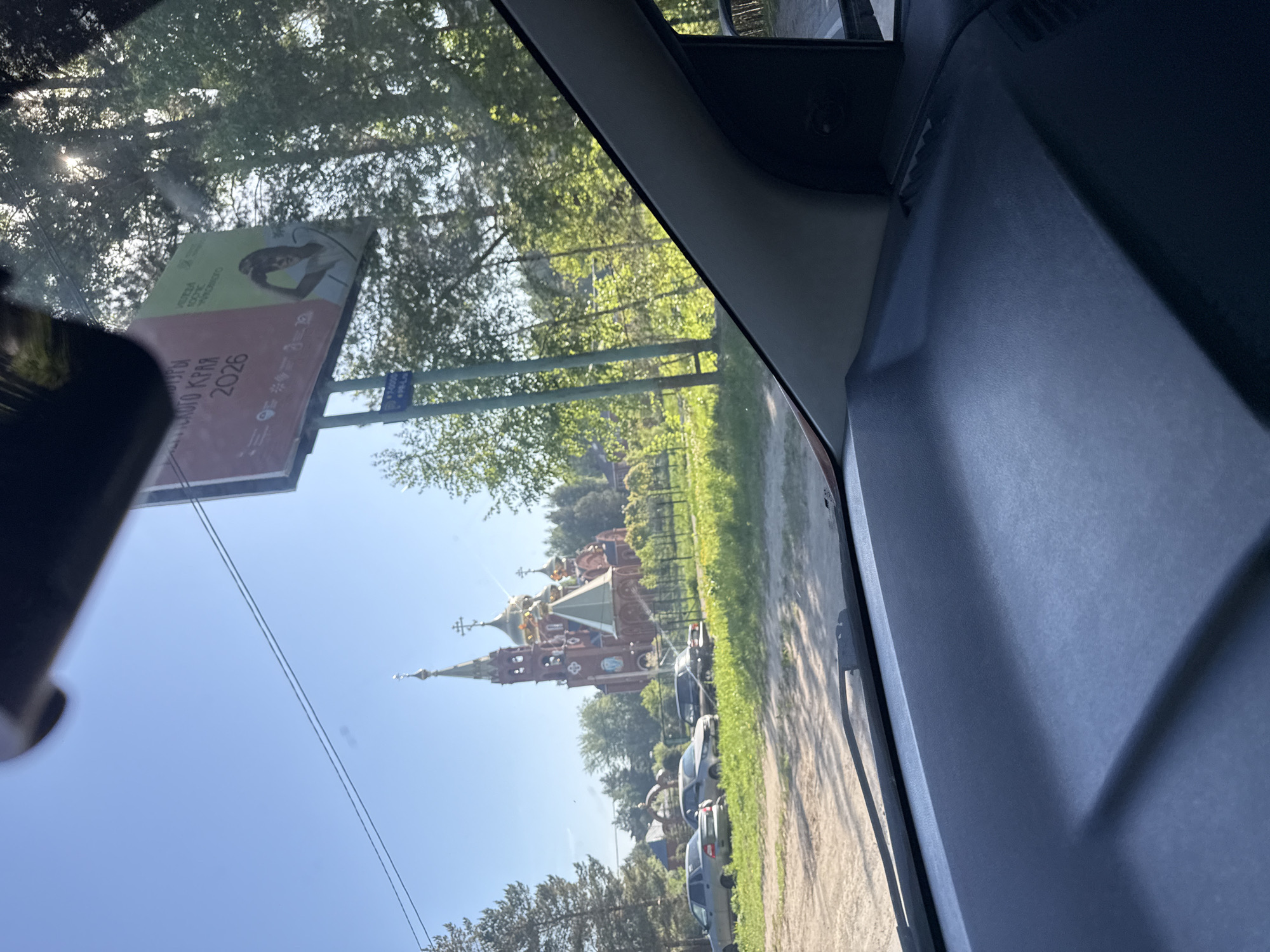 The image shows a view from inside a vehicle, likely a car. Visible through the windshield are a billboard, trees, a church with multiple domes and crosses, a road, and parked cars. The church's architecture suggests an Orthodox Christian denomination. The billboard appears to be advertising "Игры Будущего 2026", which translates to "Games of the Future 2026." Based on this information and the architecture of the church, it is most probable that the location is Chaikovsky, Russia.