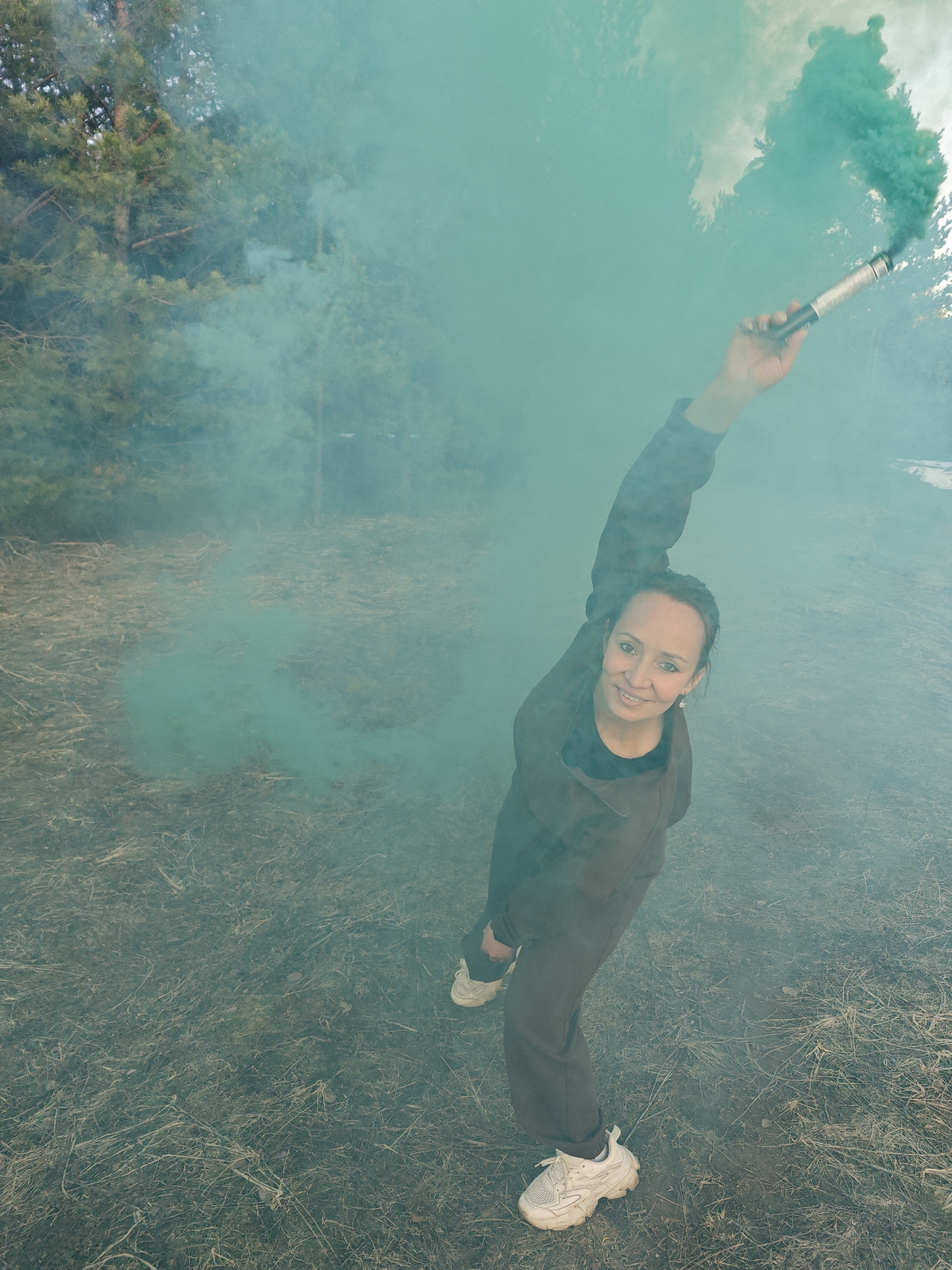 A woman is captured from a high angle in a wooded area in Dubovaya, Russia, enveloped in vibrant green or turquoise smoke. She is smiling and looking directly at the camera. She wears dark-colored clothing, possibly a tracksuit or jumpsuit, and white chunky sneakers. Her right arm is raised, holding a silver and black cylindrical device that is actively emitting the dense, colored smoke. The ground beneath her is covered in dry, light brown grass and scattered debris. In the background, green pine trees are visible, partially obscured by the swirling smoke. The scene takes place during daylight hours, creating a dramatic and somewhat ethereal atmosphere with the bright smoke. There is no visible text in the image.