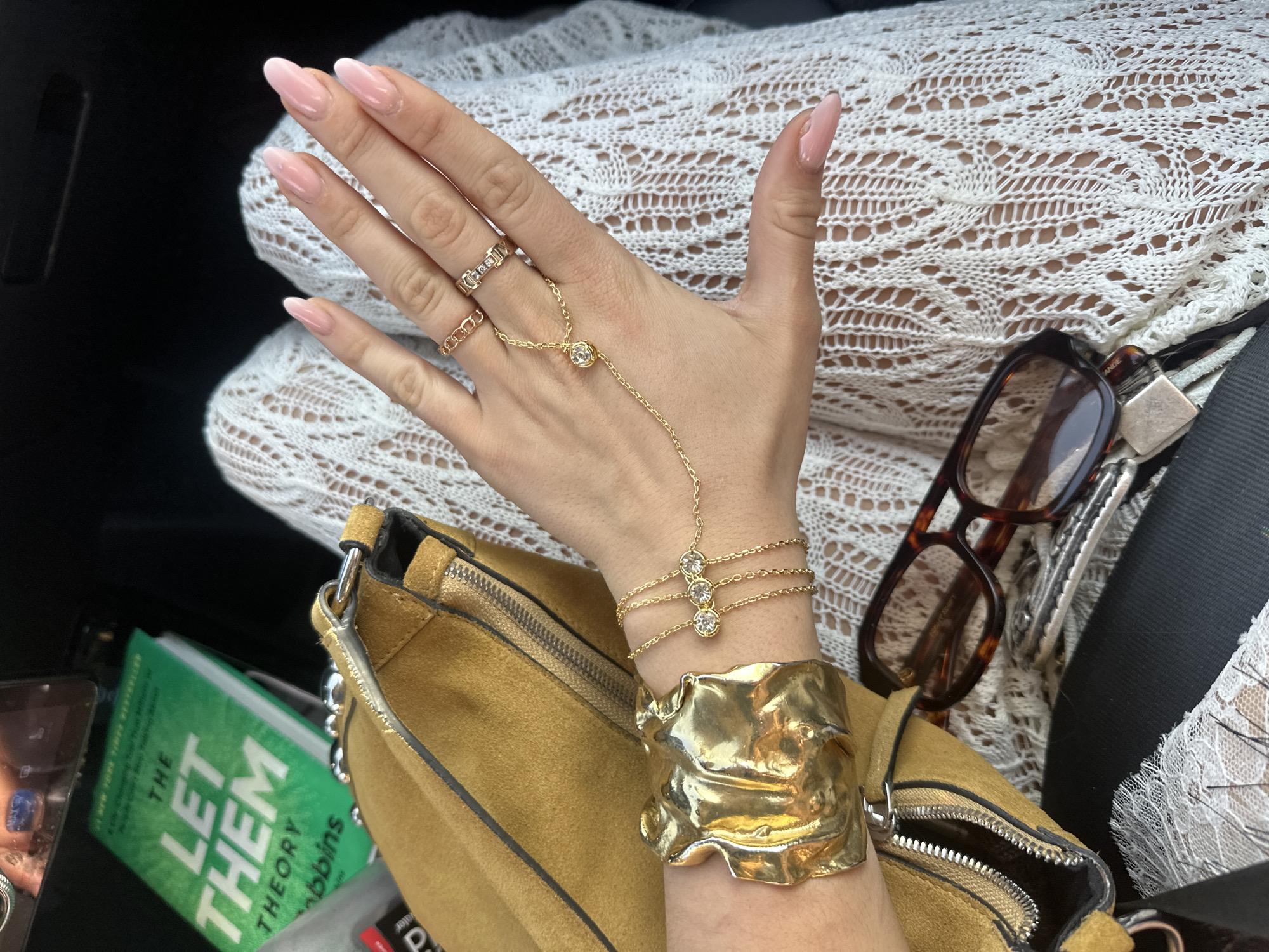 The image presents a close-up of a person's hand adorned with various jewelry pieces. Two rings are visible on the fingers, connected by a delicate gold chain to a decorative element on the back of the hand. The wrist is decorated with a multi-layered gold bracelet and a hammered, wide cuff in a yellow color. The hand rests on top of a golden suede purse. In the background, a book with green text on its cover titled "The Let Them Theory" is visible. There is also a pair of eyeglasses and what appears to be a black briefcase or bag in the scene. The location, Indio, United States of America, is not explicitly discernible from the image content itself, and is, therefore, considered extraneous information.