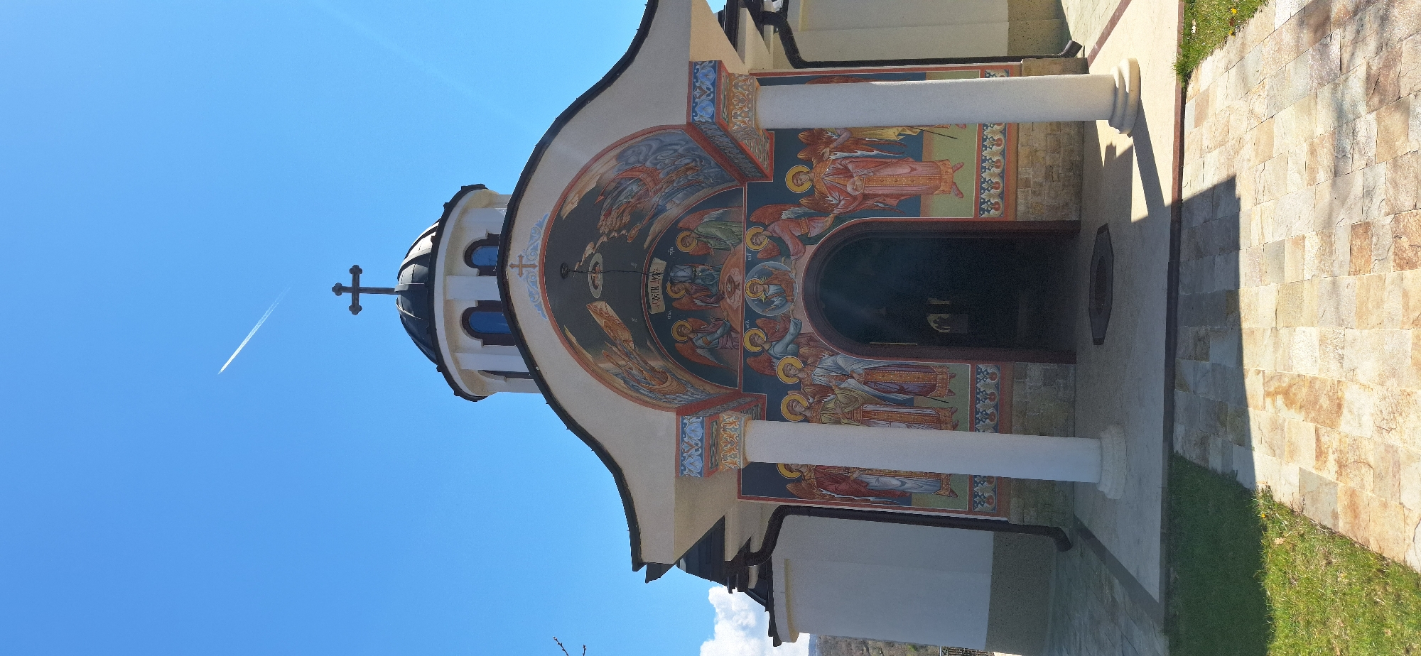 Here's a description of the image:
The image captures a section of what appears to be an Orthodox church. The building is decorated with religious iconography, depicting what appear to be angelic figures and other religious motifs painted around the entrance and under the domed roof. The entrance is framed by white columns, and there's an arched doorway leading inside. To the right of the building, there's a paved walkway or patio made of stone. The sky is clear blue with a contrail from a plane visible. The scene depicts the church located in the city of Elenovo, Bulgaria.
