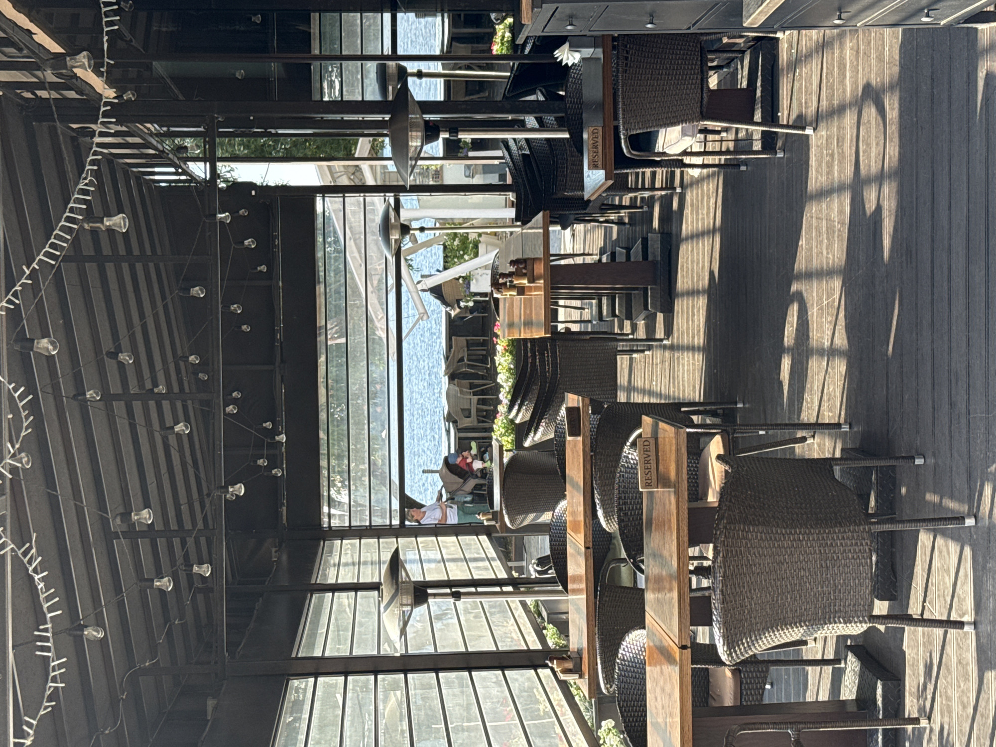 The image depicts an outdoor seating area of a restaurant or cafe with a view of a body of water. The area is covered by a roof supported by dark metal structures. String lights are hung across the ceiling. Large glass windows or panels line the sides, providing a view of the water and greenery outside. Several tables with wooden tops and wicker chairs are arranged on a wooden deck. Some tables have "RESERVED" signs. Two outdoor heaters are visible. In the background, a woman is seen pushing a baby stroller near the water. The overall atmosphere is relaxed and inviting, suggesting a pleasant dining experience with a scenic backdrop. The location is a waterfront establishment in Perm, Russia.