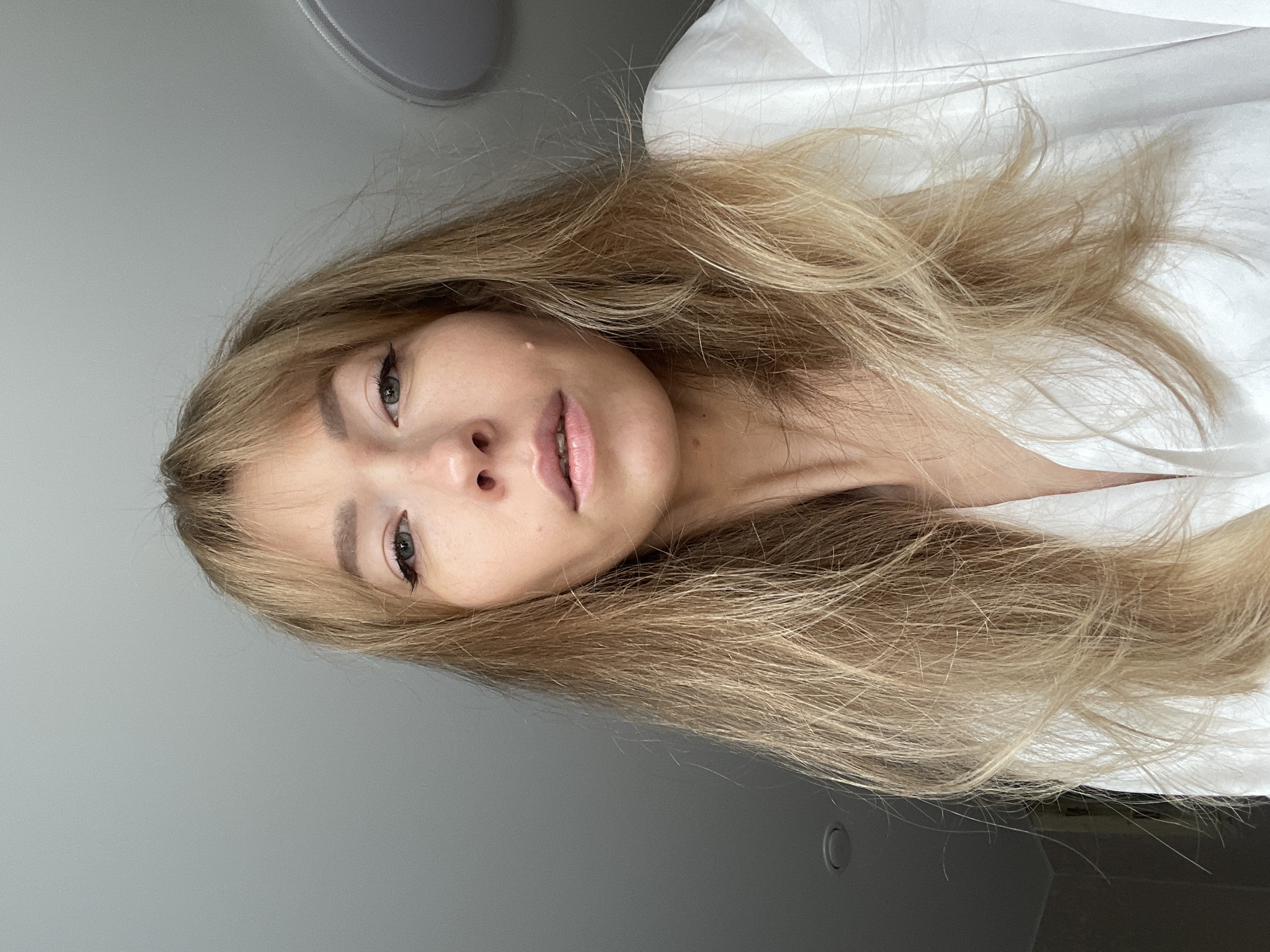 A close-up shot captures a fair-skinned woman from the chest up, with shoulder-length blonde hair framing her face. She has light eye makeup, including eyeliner and mascara, and a mole on her right cheek. The woman is wearing a white collared shirt, partially unbuttoned. The background is a plain, light gray wall with a circular light fixture visible in the upper right corner. The image appears to be taken indoors. There is no information to indicate the location is in Perm, Russia.