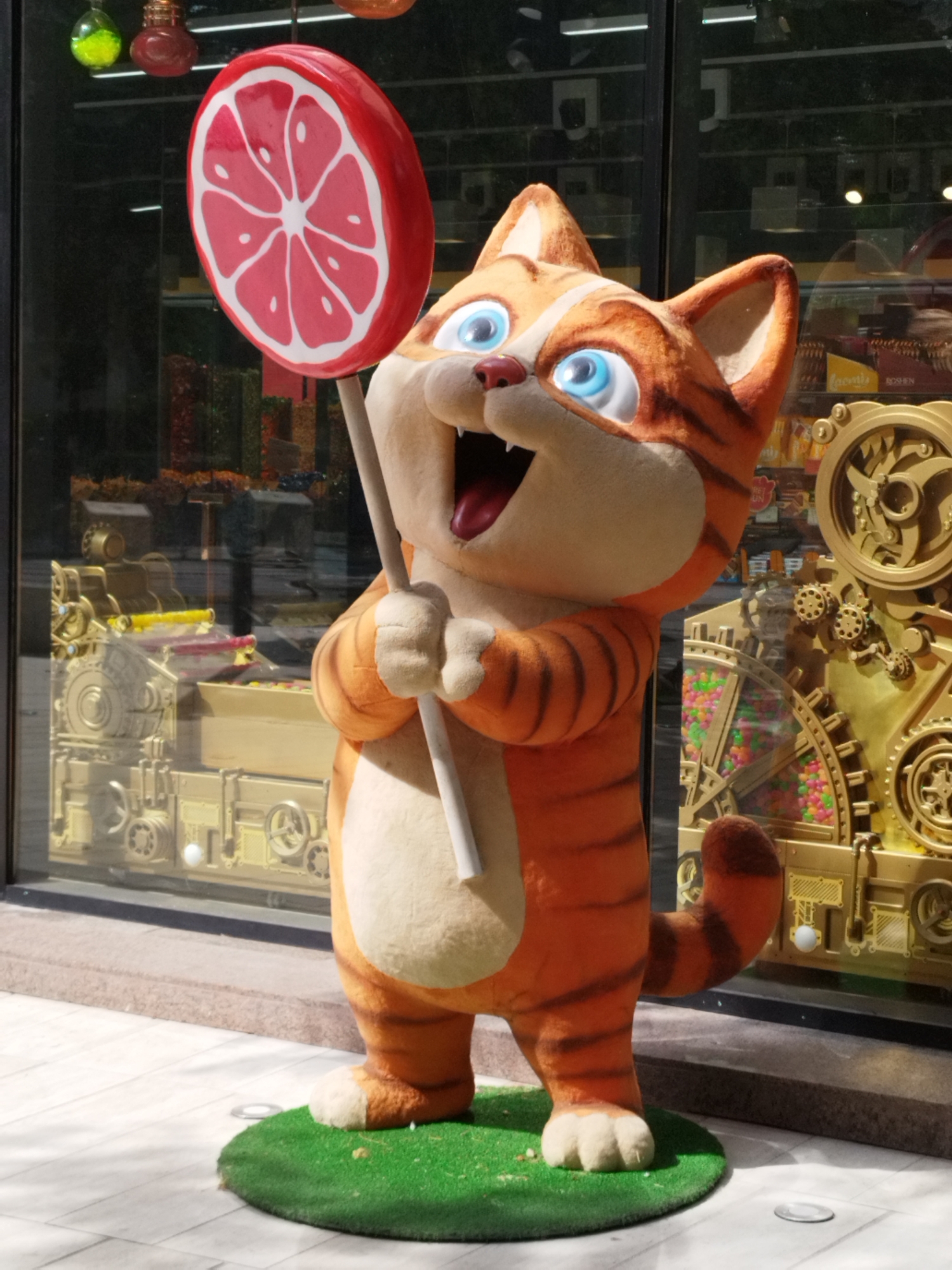 An outdoor statue of a cartoon cat, likely a promotional display, stands on a patch of artificial grass. The cat is orange with black stripes and has large blue eyes and an open mouth with a red tongue visible. It is holding a large, red and white lollipop that resembles a slice of grapefruit on a stick. Behind the statue is a shop window displaying various items, including what appear to be candies and a golden, intricate mechanical-looking structure. The ground in front of the statue is paved with light-colored tiles, and sunlight casts shadows. The scene appears to be located in an urban setting, possibly a commercial street or a shopping area.