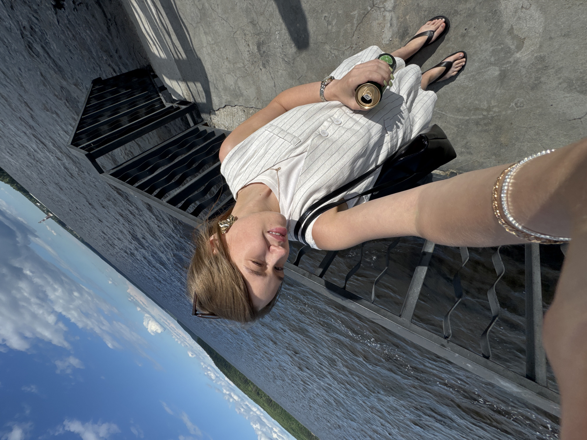 A woman takes a selfie on a sunny day next to a body of water. She is wearing a white pinstripe vest over a white t-shirt, black flip-flops, and a gold bracelet. She is holding a can of what appears to be a beverage in her right hand. In the background, a black metal railing lines the edge of a concrete embankment, and a green, tree-covered shoreline is visible across the water. The sky is blue with scattered white clouds. The location is the embankment of the Kama River in Chaikovsky, Russia.