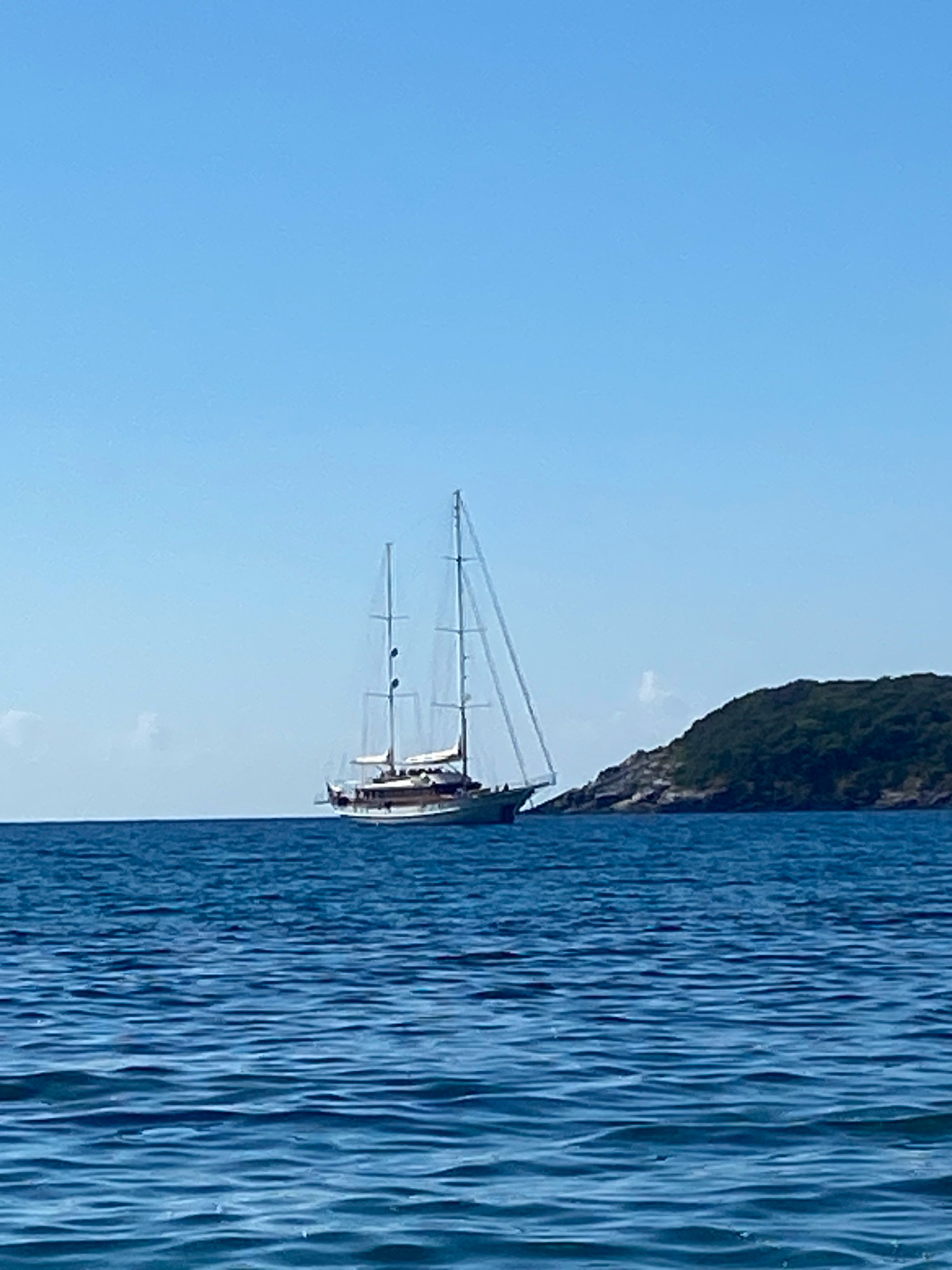 A wide shot captures a tranquil blue sea under a clear, pale blue sky. A large, wooden sailing vessel with two prominent masts and several sails, some furled, cruises on the calm water. To the right of the sailboat, a verdant, tree-covered hill rises from the sea. The scene is bathed in sunlight, creating a serene and idyllic atmosphere. The location is Prijevor, Montenegro.