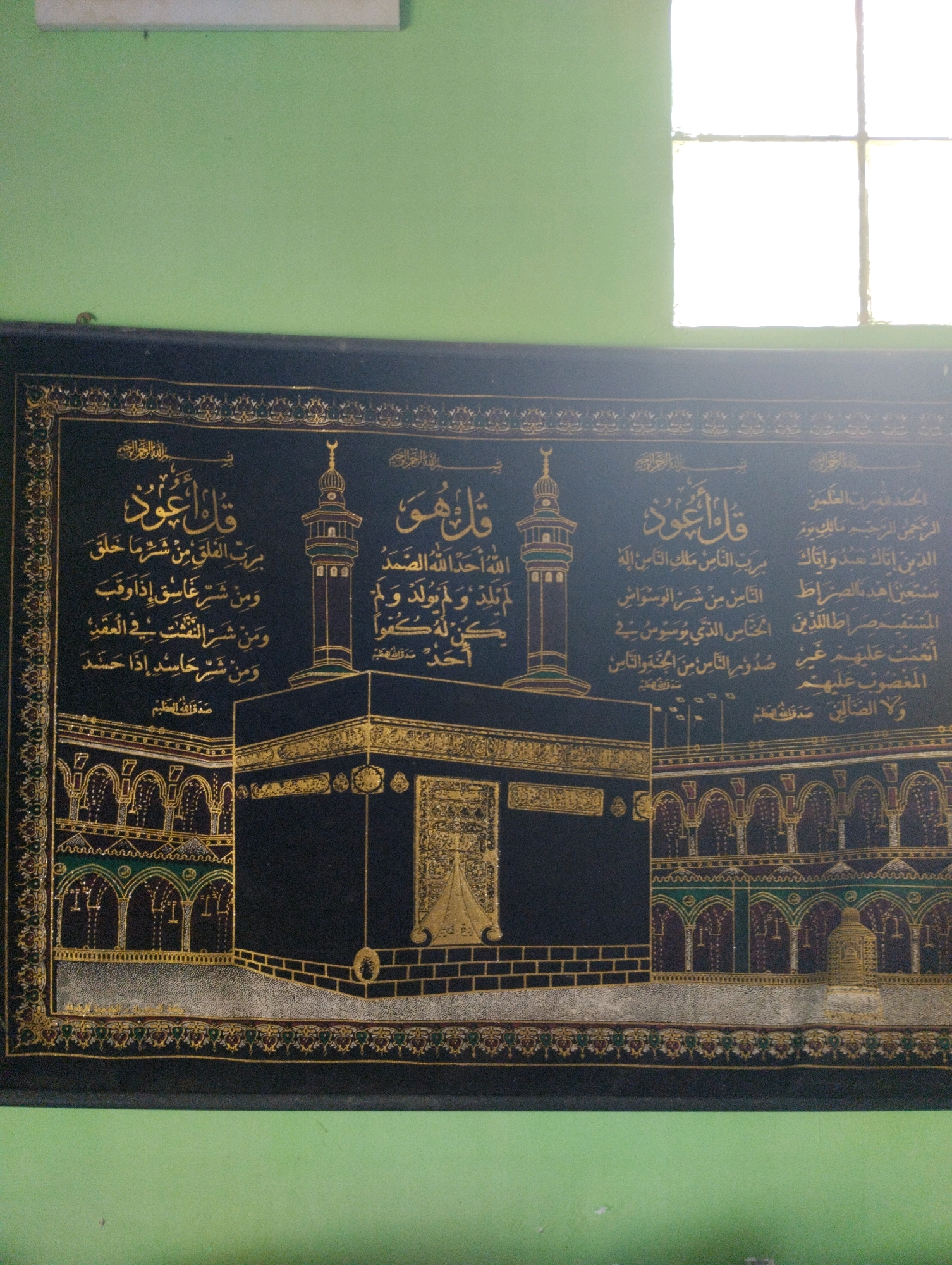 The image shows a tapestry depicting the Kaaba in Mecca. The tapestry is black with intricate gold embroidery featuring Arabic calligraphy and architectural details of the Kaaba and its surrounding structures. The tapestry is hanging on a light green wall, partially illuminated by natural light from a window on the right. The scene appears to be indoors. The prompt claims the image is located in the city of Cimenyan, Indonesia, though there is no visual evidence in the image to support this specific location.