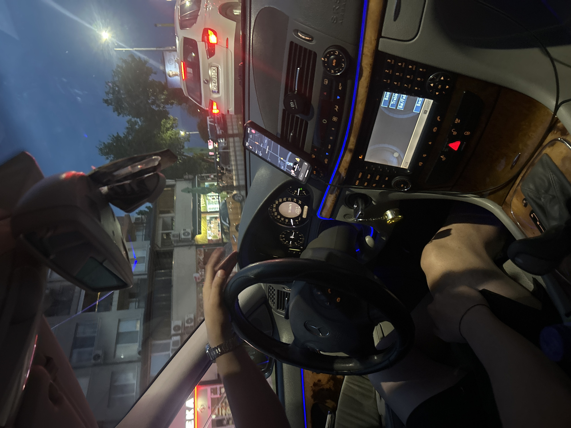 The scene depicts an urban environment in Plovdiv, Bulgaria, observed from the driver's perspective inside a Mercedes-Benz vehicle at dusk or night. The interior features a three-spoke steering wheel with the Mercedes-Benz emblem, a dashboard with visible instrument cluster indicating a speed of 16 km/h, an odometer reading of 270,776.0 km, and an ambient temperature of +27.5°C. The central console, adorned with wood grain trim, includes a display showing audio settings such as "Bass," "Treble," and "Aux." The hazard warning lights are illuminated. A smartphone, displaying a map application, is mounted on a magnetic holder positioned on the right-side air vent. Blue ambient lighting illuminates the lower dashboard and door trim. A keychain with a golden charm hangs near the gear shifter. The driver's hands are visible, with the left hand wearing a metallic watch and a black wristband resting on the steering wheel, and the right hand resting on top of the steering wheel. A second, bare hand, also with a black wristband, is visible resting on the driver's upper thigh, suggesting an interaction between the driver and a passenger.
Outside the vehicle, a white BMW X3 with Bulgarian license plate "CB 4733 CK" is directly in front, stationary in traffic. The street is illuminated by streetlights and the taillights of other vehicles. Buildings with multiple stories and visible air conditioning units line both sides of the street. Storefronts with illuminated signs, including a red sign partially showing "EKA," are visible on the left. Trees are silhouetted against the dark blue sky, and a distant billboard features a man's face.