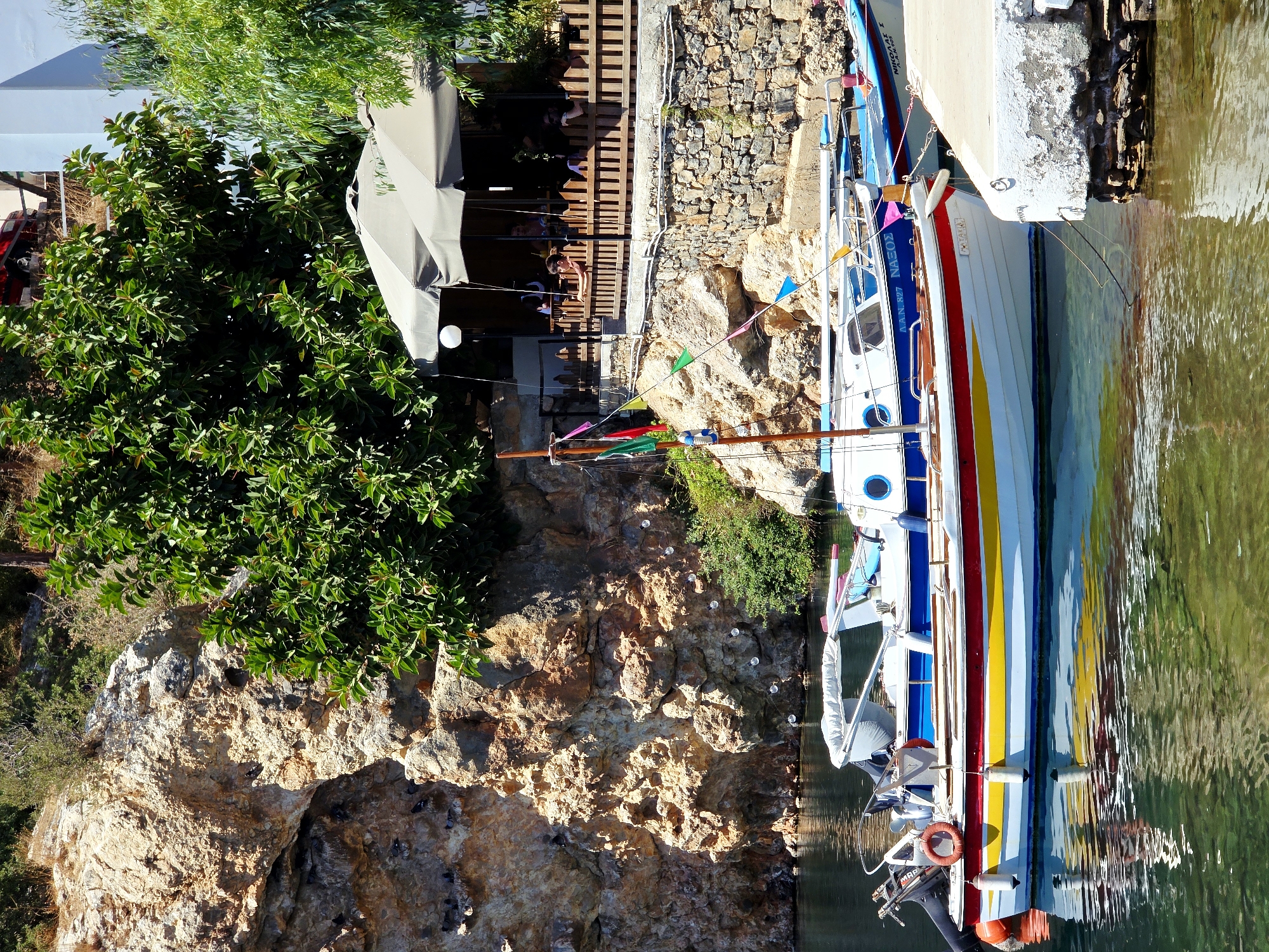 The image depicts a waterfront scene in Agios Nikolaos Municipal Unit, Greece. In the foreground, two boats are moored in clear, green water. The closest boat is a white vessel with red, yellow, and blue stripes along its hull. It has a white canopy and several fenders. The second boat, behind the first, is primarily blue and white. In the middle ground, a stone wall runs along the shoreline, topped by a wooden fence. Behind the fence, a shaded outdoor seating area of a restaurant is visible, with several people seated at tables under a large, cream-colored awning. A lush green tree with large leaves overhangs the restaurant. The background features a steep, rocky cliff face with sparse vegetation. Sunlight illuminates the scene, casting shadows and highlighting the textures of the rock and water.
