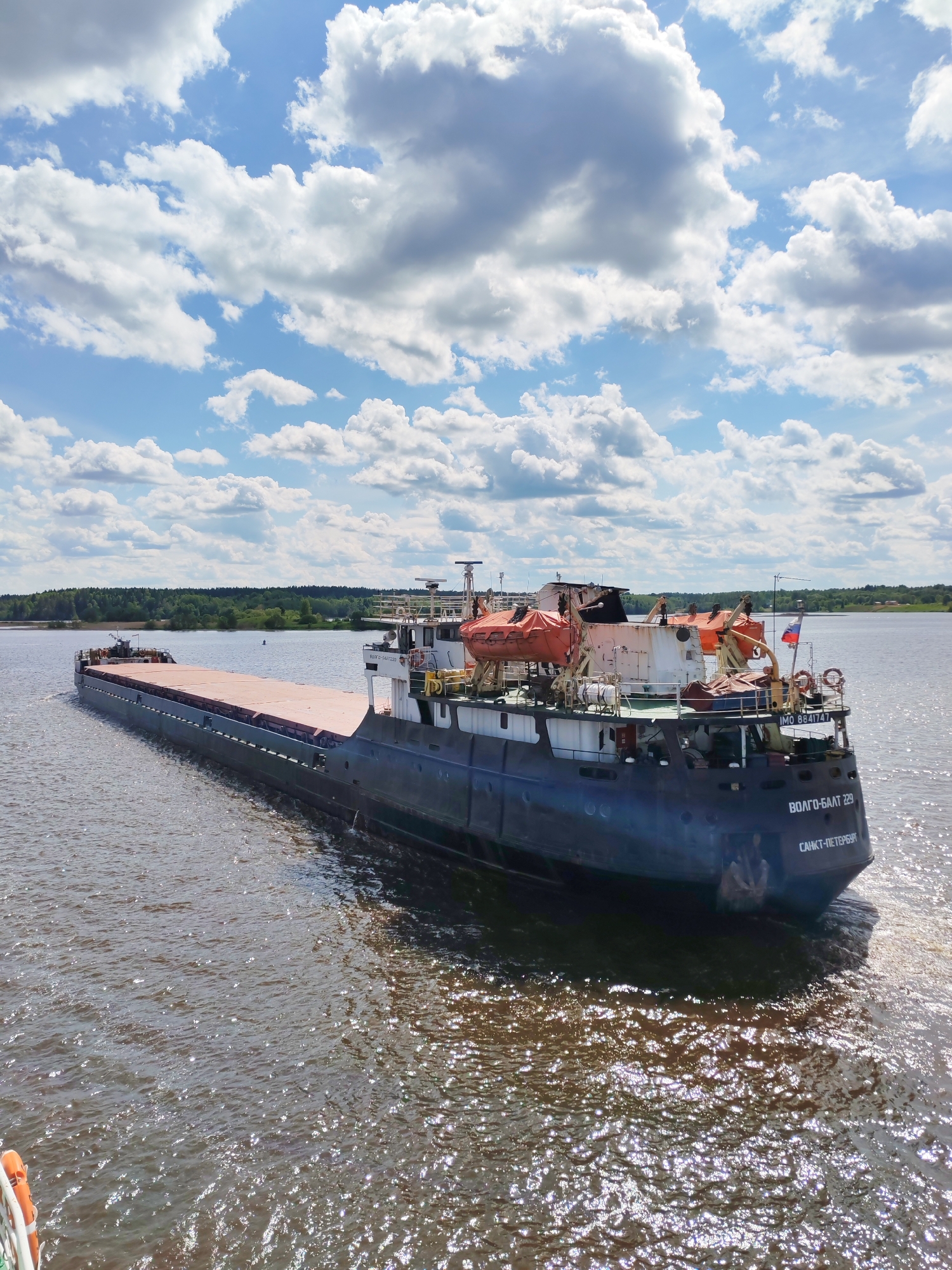 A large cargo ship, identified as "VOLGO-BALT 229" and registered in "CAHKT-ПETEPБYPT" (Saint Petersburg), is seen navigating a body of water in Goritsy, Russia. The vessel is mainly dark blue, with a flat, reddish-brown cargo deck. At the stern, there are visible lifeboats, a Russian flag, and the ship's IMO number "IMO 8841747." The water has a slightly choppy surface with sunlight reflecting off the waves. The horizon displays a green shoreline of trees. The sky is partly cloudy with patches of blue. The photo seems to be taken from another boat or platform, as part of a railing is visible in the lower left corner.