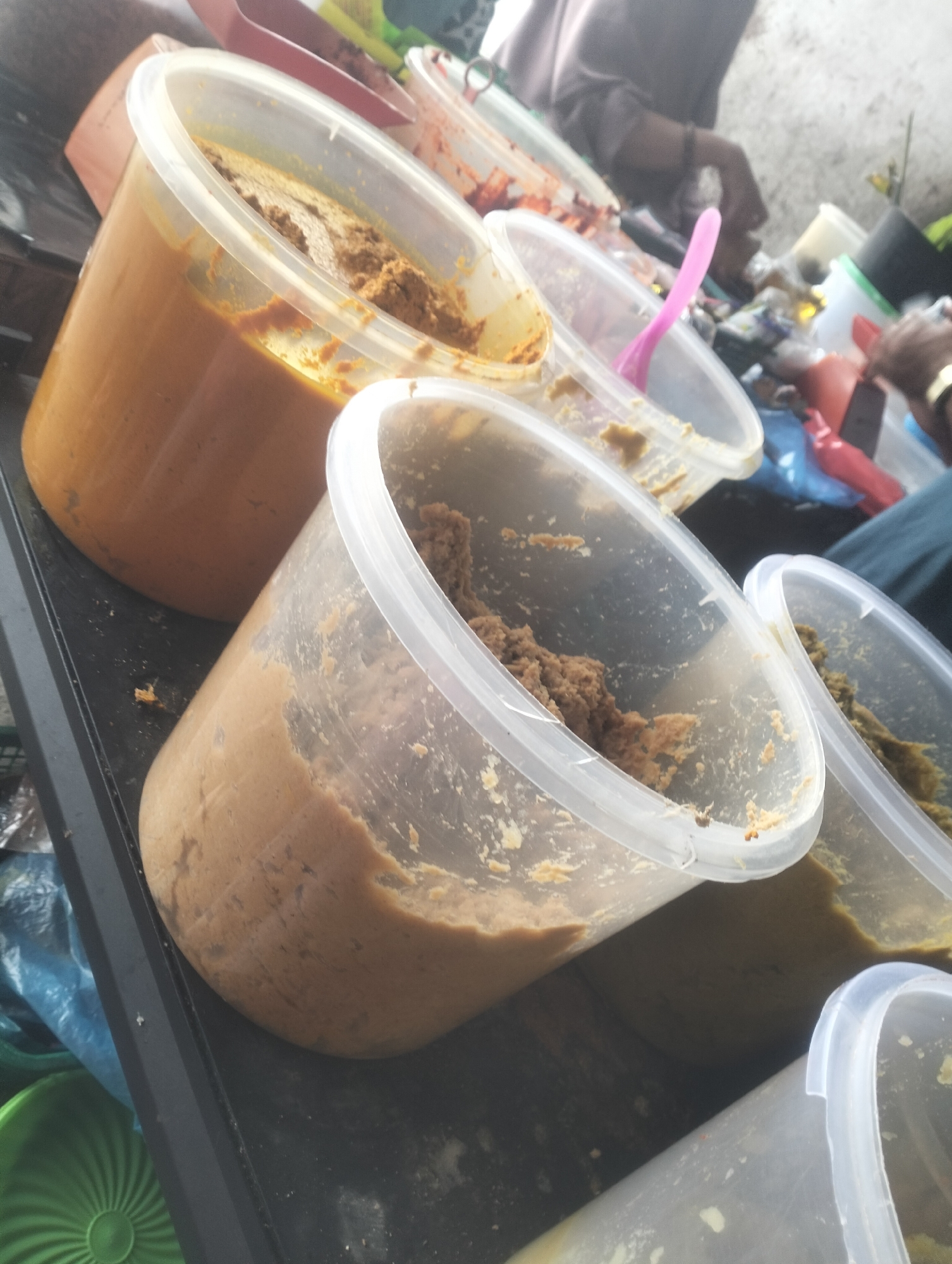 In the city of Dumai, Indonesia, a market scene unfolds with a focus on several large, clear plastic containers filled with various food items. The largest container, positioned prominently on the left, holds a thick, orange-brown paste, possibly a curry or sauce. Adjacent to it, another container contains a similar substance, appearing slightly darker and more textured. A third container to the right holds a lighter brown, paste-like food, with visible flecks. Further to the right, another container, partially visible, appears to contain a green food item. The containers are arranged on a dark surface, likely a table or stall. In the background, a person's arm, wearing a watch, is visible reaching towards one of the containers. Another person's torso, in a muted pink or purple garment, is also visible in the upper background, suggesting human interaction within the market environment. The overall setting appears to be an outdoor or semi-enclosed market space, with hints of a wall and other market goods in the blurry background. The lighting suggests an indoor or shaded area, as there are no strong direct shadows.