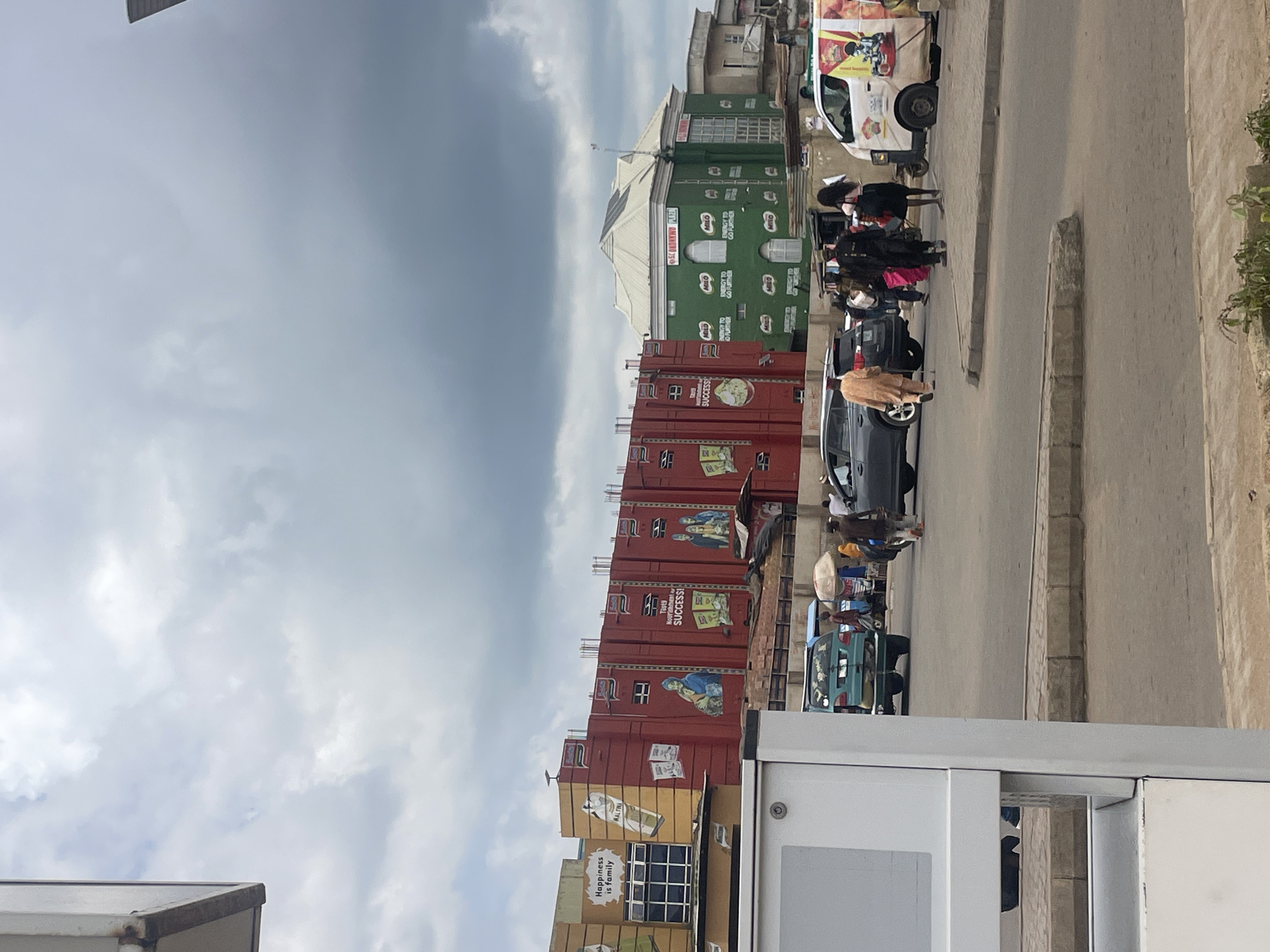 This image captures a street scene in Jos, Nigeria, under an overcast sky. In the background, a large, red building is adorned with advertising murals, one prominently featuring the slogan "Happiness is family" and another depicting images related to "SUCCESS." Adjacent to the red building is a smaller, green structure with "S.C OKONKWO PLAZA" visible on its facade, also covered in advertisements for "MILO" with the tagline "ENERGY TO GO FURTHER."
In the foreground and middle ground, a street intersection is active with pedestrian and vehicular traffic. Several people are walking, including one individual in an orange robe and another in a dark uniform who appears to be directing traffic or observing. Vehicles, including a blue SUV, a lighter colored SUV, and a white van with colorful advertisements, are present on the road. A man with a white umbrella walks beside a cart. The road surface appears to be asphalt, with concrete curbs and sidewalks. A white, partially visible structure, possibly a kiosk or a service point, is in the left foreground. The environment suggests a commercial or bustling urban area.
