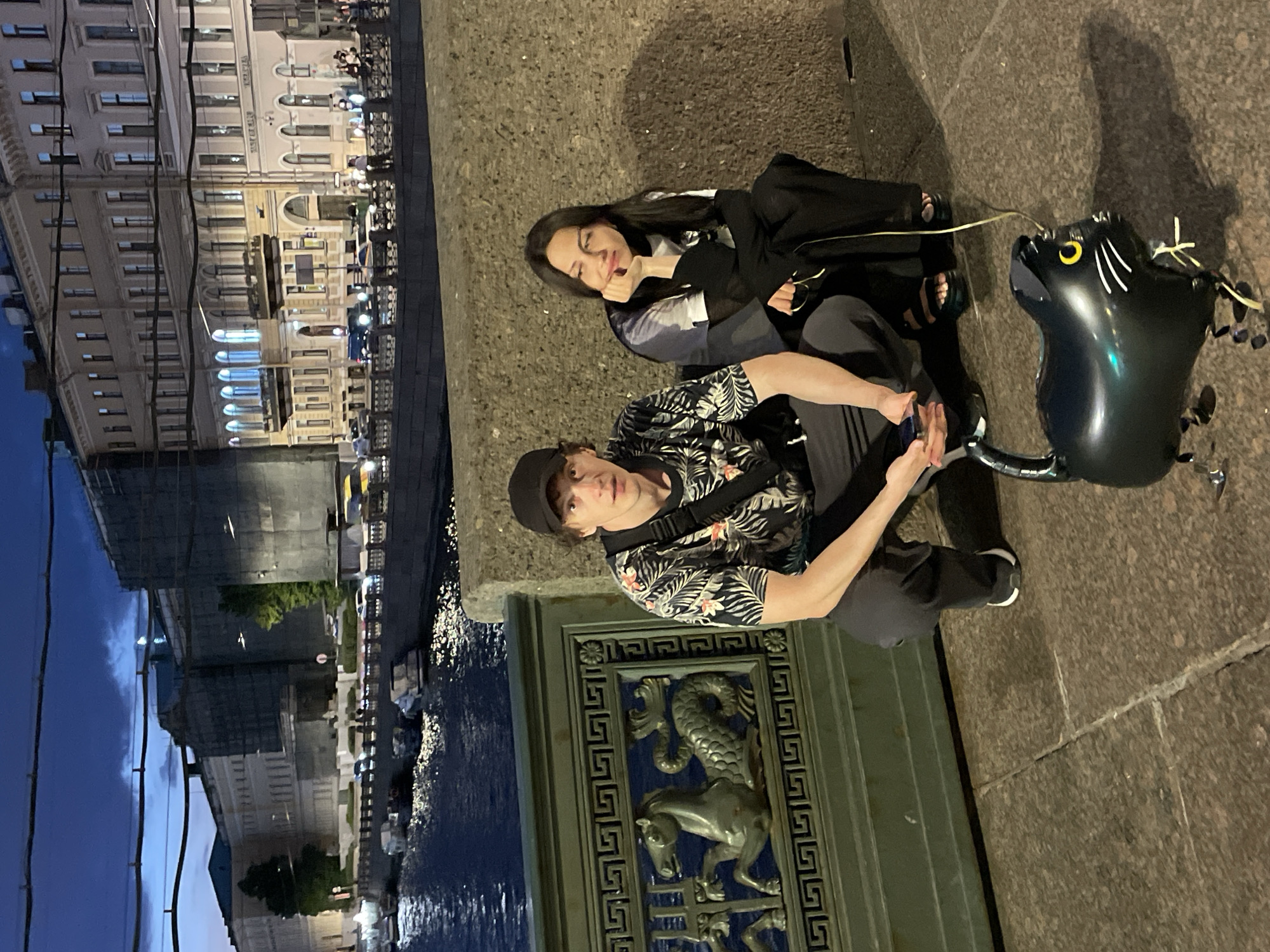Two individuals, a man and a woman, are seated on a stone ledge in front of a decorative metal railing along a body of water. The man, wearing a black baseball cap and a patterned short-sleeved shirt, is crouched down, holding a smartphone. The woman, with dark hair, is seated next to him, resting her chin on her hand. A black cat-shaped balloon, tethered by a yellow string, is positioned on the ground in front of them. In the background, a river is visible, with boats and buildings lining the opposite bank. The architecture suggests a European city, and the style of the buildings, particularly the classical elements and the waterfront setting, are characteristic of Saint Petersburg, Russia. The time appears to be dusk or early evening, as evidenced by the subdued lighting and the illuminated windows of the buildings.