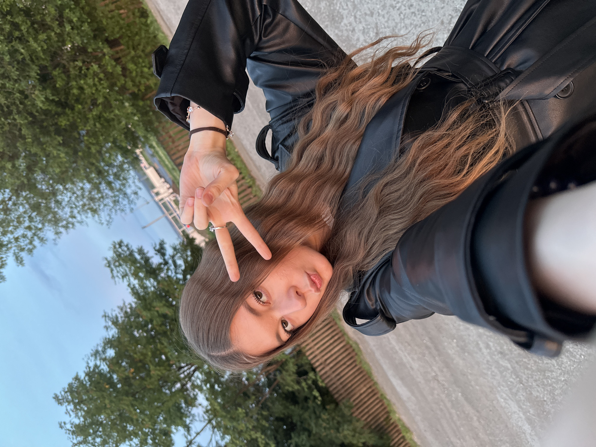 A young woman is holding up two fingers in a "peace sign" in front of her face, while wearing a black leather jacket. Her long, wavy, brown hair is hanging down over her jacket. In the background, there are green trees and a blue sky. The woman is standing on a paved surface. There is no indication of the location being in the city of Кузембетьево, Russia.