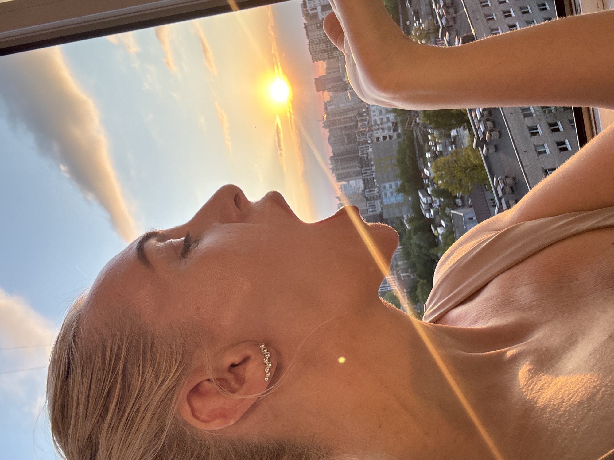 A low-angle, medium shot captures a woman looking up at the sun setting behind high-rise buildings. She has fair skin, blonde hair, and an ear cuff with a row of small stones. Her mouth is slightly open, and her face is angled towards the sky. A portion of her arm is visible, positioned upwards as if reaching toward the sun. The background shows a cityscape with tall buildings and some greenery. The setting sun casts a warm glow on the woman and the surrounding environment.