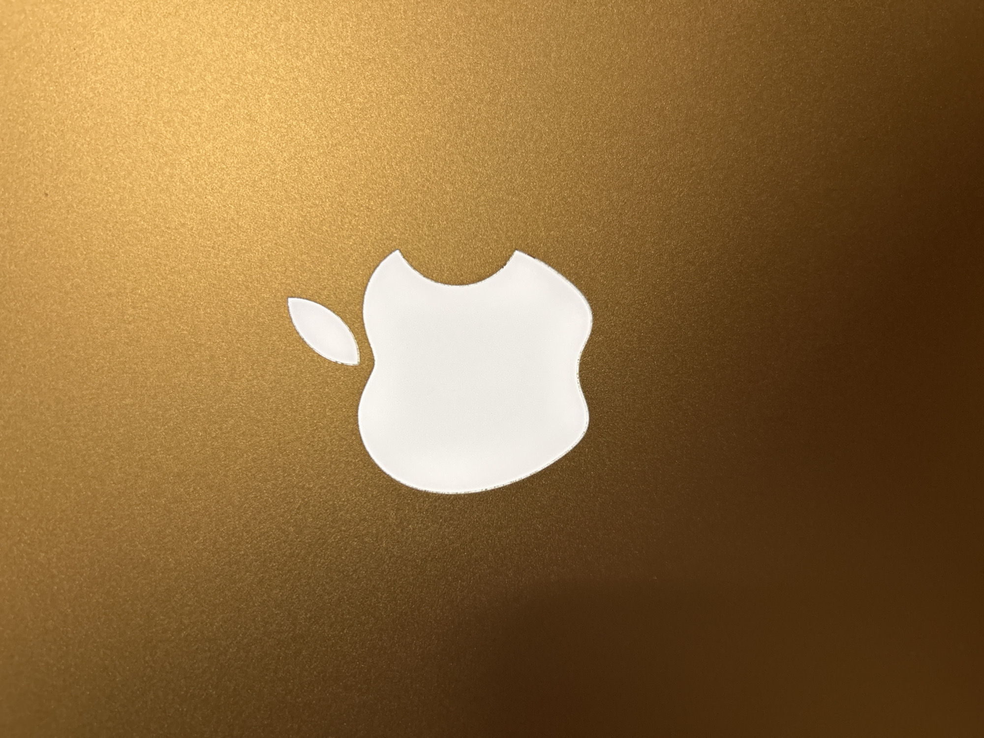 This media file is a close-up, eye-level shot of the Apple logo, which is a white silhouette of an apple with a bite taken out of it. The logo is centered on a textured, gold-colored surface, likely the casing of an Apple device. The lighting suggests a bright, possibly indoor environment, casting subtle shadows and highlighting the grain of the gold material. There is no visible text in the image. The scene is static, focusing solely on the iconic branding against its metallic backdrop. The image appears to be taken in Kadewatta, Sri Lanka, though this context does not directly influence the visual content of the logo itself.