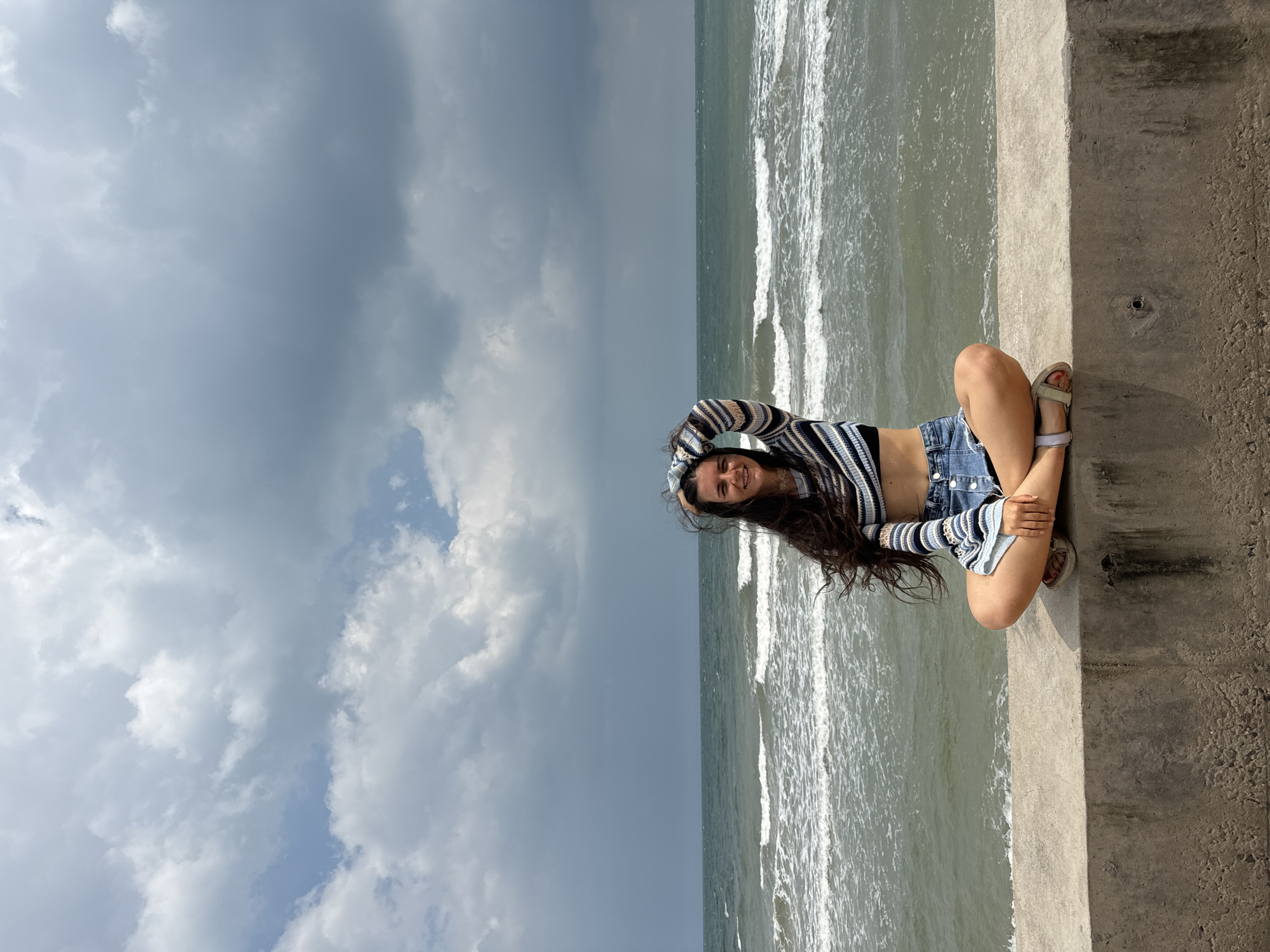 A young woman sits cross-legged on a concrete barrier facing the sea. She has long, dark hair that is being blown by the wind. She is wearing a striped cropped sweater and denim shorts. The sea behind her is choppy, with white waves. The sky is cloudy, with some patches of blue visible. The location is the coast of Phu Thuy Ward, Vietnam.