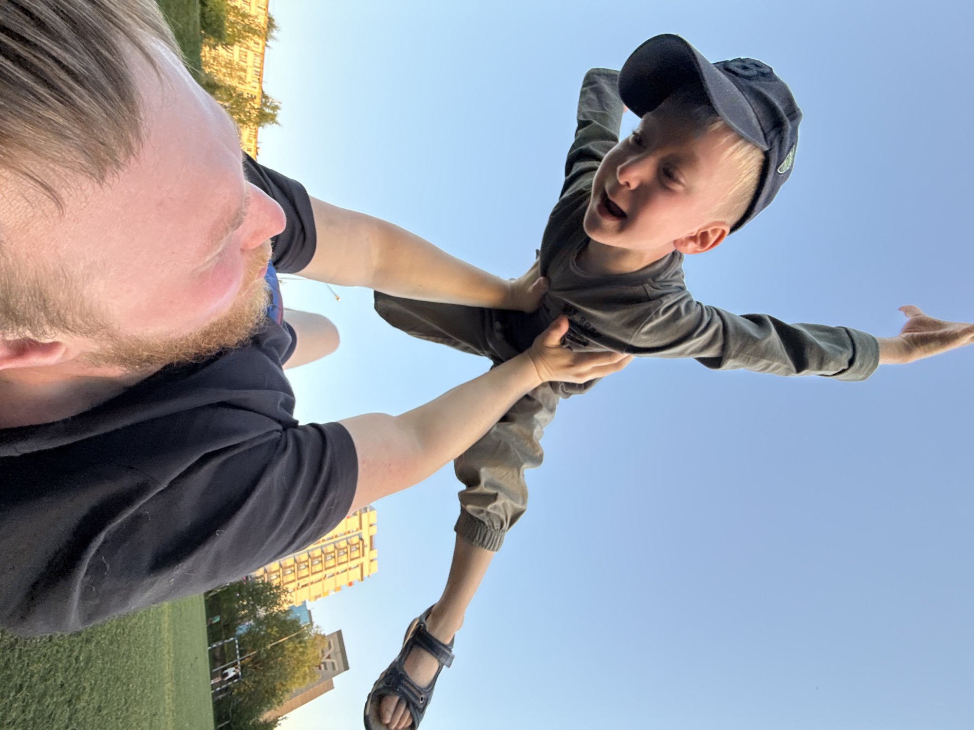 A man with light brown hair and a beard is holding a young boy in the air. The boy is wearing a baseball cap, a long-sleeved shirt, pants, and sandals. The scene appears to be outdoors, possibly in a park or green space in the city of Chaikovsky, Russia, with buildings and trees in the background and a clear blue sky above. The boy's arms are outstretched, as if he is flying.