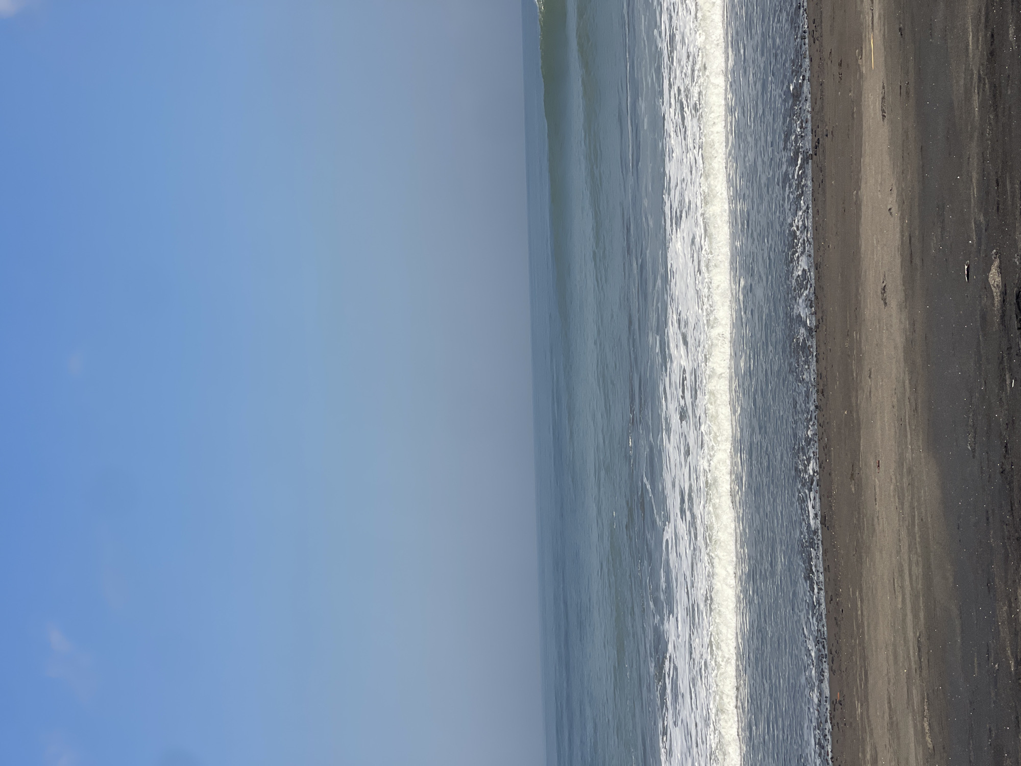 The image shows a view of the ocean and beach under a clear blue sky. The foreground is a dark, sandy beach with small pebbles visible near the water's edge. A wave is breaking in the mid-ground, with white foam spreading across the dark water. The ocean extends to the horizon, where it meets a hazy, light blue sky. There are no people or man-made objects visible in the image. The scene appears to be a natural coastal environment. The mention of Kediri, Indonesia is not supported by any visual information in the image.