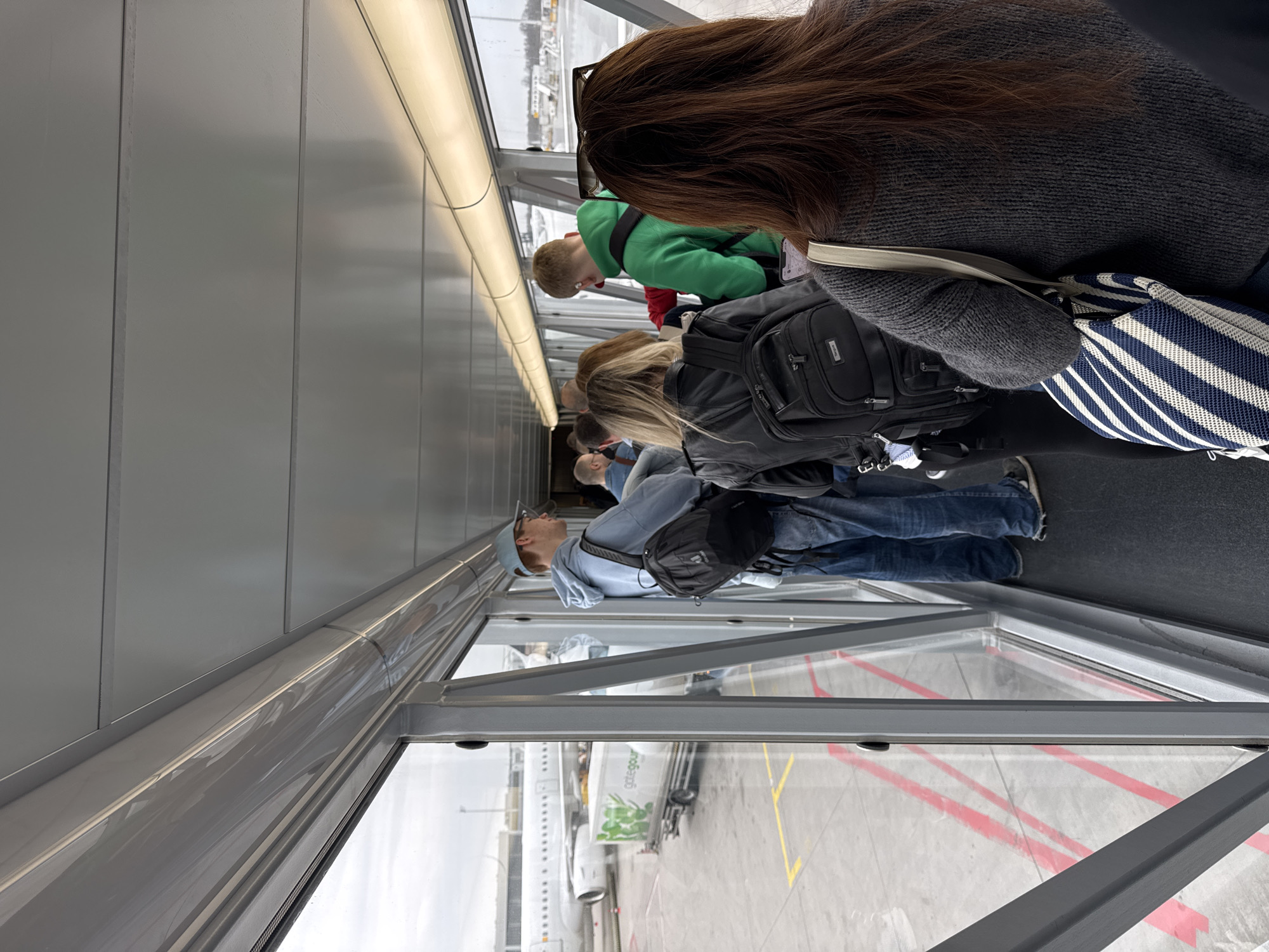 The photograph was taken in Oberding, Germany, inside an airport jet bridge. The image shows a line of passengers boarding or disembarking an airplane, likely a Gate Gourmet plane based on what can be seen outside the window. Passengers are wearing a variety of clothing. The windows of the jet bridge are visible, providing a view of the tarmac. The jet bridge is lined with gray panels. The image is taken from the perspective of someone standing in the line of passengers.