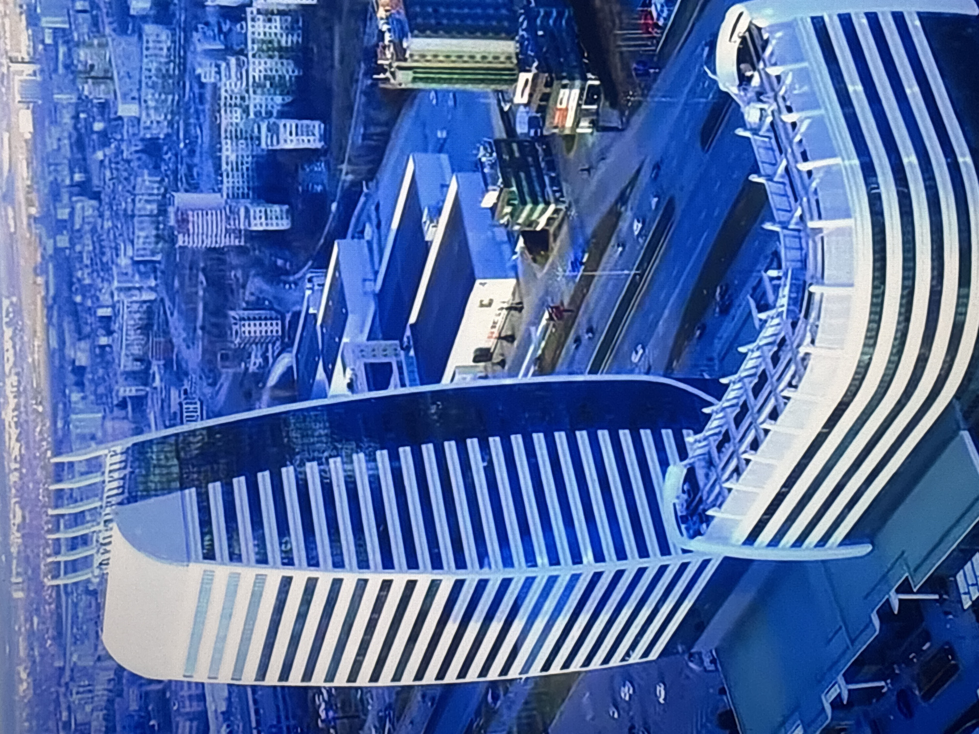 This aerial photograph depicts a bird's-eye view of a contemporary, curved building in Sofia, Bulgaria. The building features a distinctive striped pattern of white and dark-colored vertical lines across its facade. The structure has a curved design with a modern architectural style. The surrounding environment shows a mix of buildings of varying sizes and designs, along with roads. The image is mostly blue tinted.
