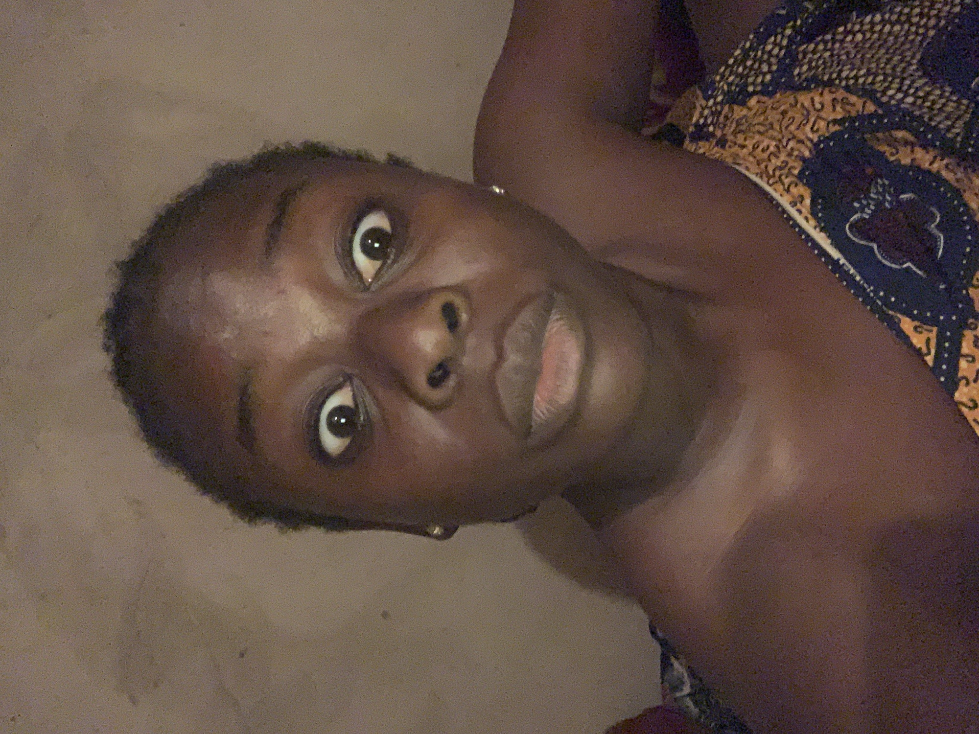 The image shows a close-up, selfie-style portrait of a young Black woman with her eyes wide open, appearing surprised or startled. She is wearing a patterned garment that covers her right shoulder and chest. The background appears to be a plain, textured wall. The lighting is dim, suggesting an indoor setting at night. The woman's facial expression is the focal point, with her pupils dilated and her mouth slightly open. No other individuals or significant objects are visible in the frame. The image does not contain any information indicating its location in Agban, Nigeria, or any other specific geographical area.
