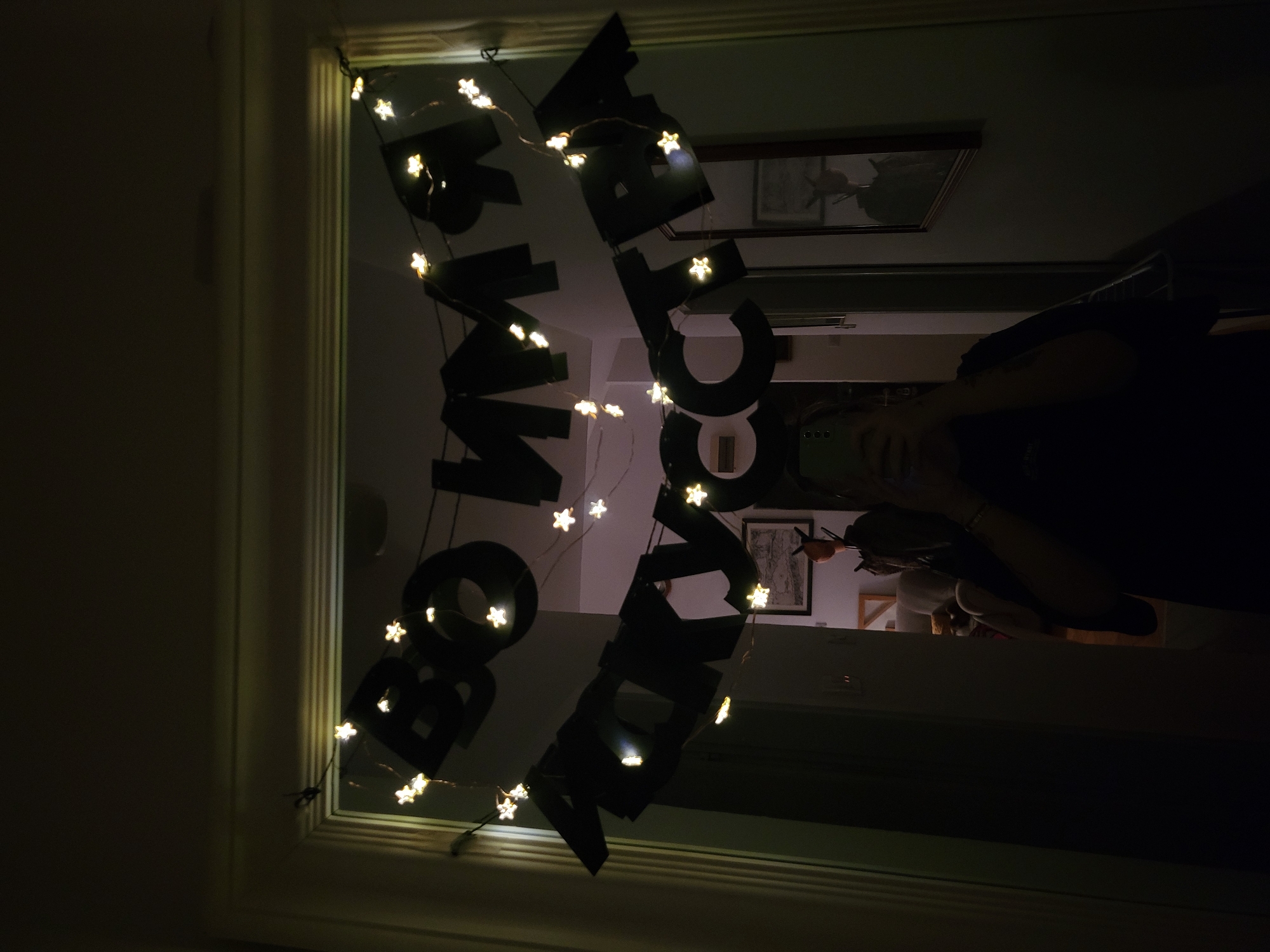 The image shows a decorated doorway with the words "BOO HAPPY HALLOWEEN" spelled out in large, black, block letters. The letters are strung across the doorway and intertwined with a string of small, star-shaped fairy lights. The doorway itself is white and rectangular. The view through the doorway reveals a glimpse of the interior, including framed artwork on the wall and a person holding a mobile phone. Given the decorations, the context suggests it's a Halloween celebration. While the image does not provide sufficient information to confirm the location as Budva, Montenegro, the interior appears to be an apartment hallway.