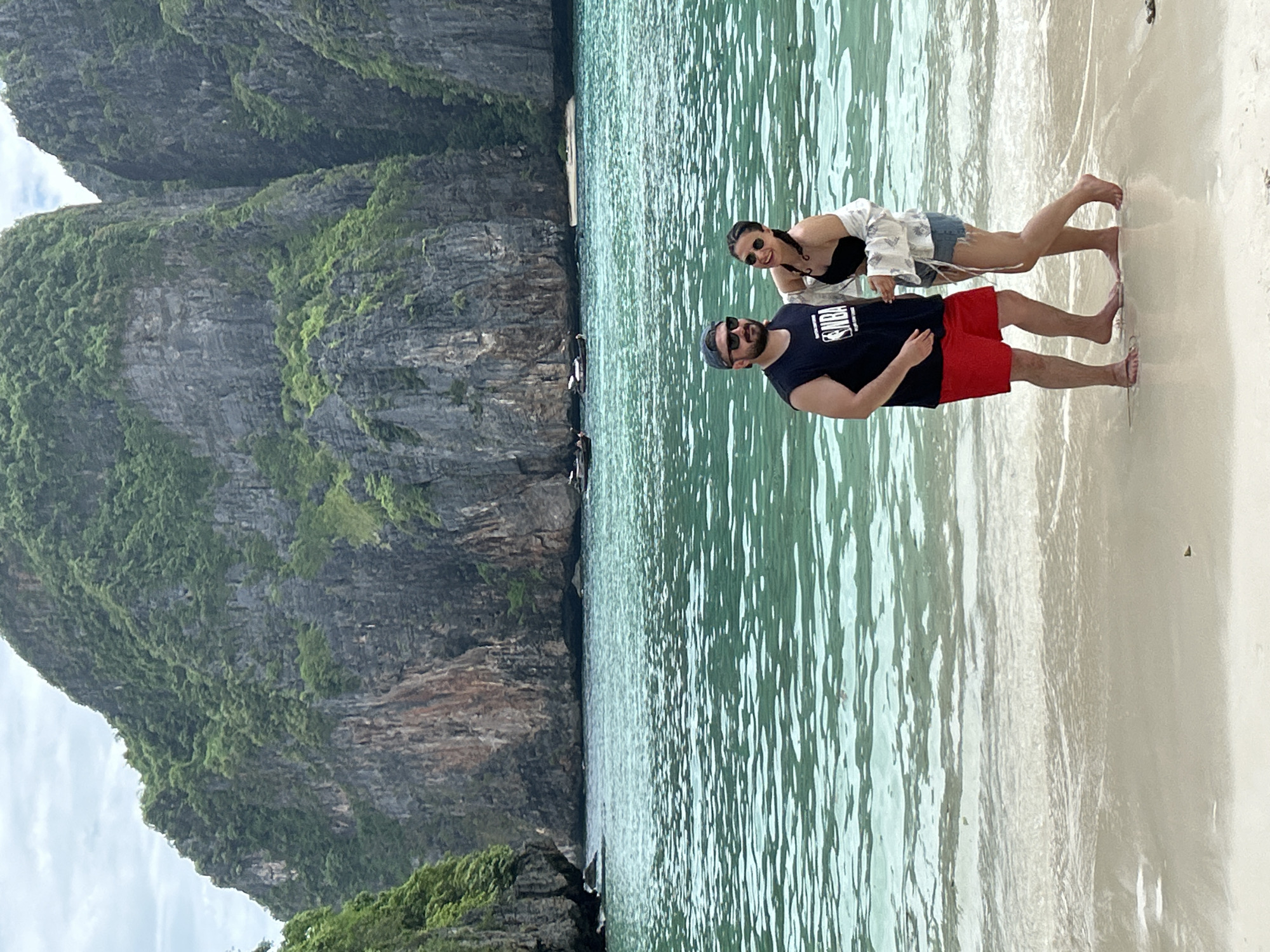 An outdoor, medium-shot photograph captures a couple standing on a sandy beach at the edge of turquoise water. The man, on the left, wears a sleeveless black t-shirt with "NBA" logo, red shorts, a blue baseball cap, and sunglasses. The woman, on the right, is dressed in a black bikini top, denim shorts, and a light-colored, patterned overshirt. They stand close together, with the man's arm around the woman's shoulders and her hand on his chest. They are both smiling.
In the background, a body of water with gentle waves extends to large, imposing limestone cliffs covered in lush green vegetation. Several longtail boats are visible in the water near the cliffs. The sky above is overcast with clouds. The overall scene depicts a tropical beach setting, likely in Ao Nang, Thailand, known for its dramatic karst formations and clear waters.