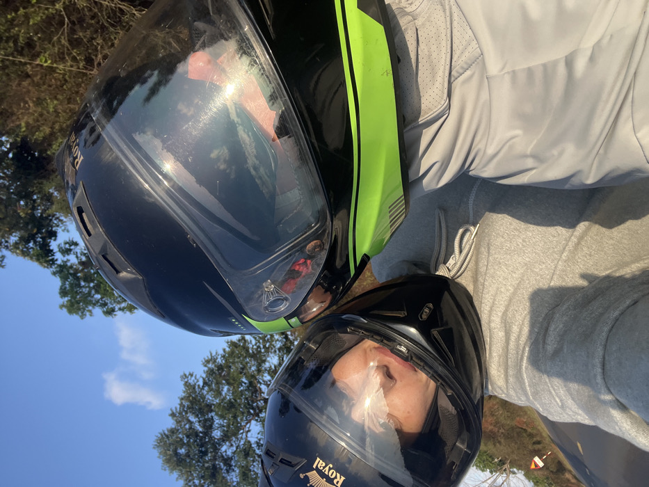 This image depicts two individuals wearing motorcycle helmets, positioned closely together in an outdoor setting reportedly in Ea Ksô, Vietnam. The individual on the left wears a black full-face helmet with "Royal" branding and a gold crown logo, a clear visor partially reflecting the sky. Their face is visible behind the visor, with white tissue placed on their nose. This individual is wearing a light grey hooded sweatshirt with visible white drawstrings. The individual on the right wears a black full-face helmet featuring prominent neon green stripes and accents, with a clear visor also showing reflections of the sky and light sources. Their face is obscured. This individual wears a light grey garment, possibly a jacket or shirt, exhibiting perforated texture details. Both individuals' helmets are positioned with their visors nearly touching. The background features a clear blue sky with a few wispy clouds and a line of green and reddish-brown trees. A segment of a dark asphalt road and a partially visible white and orange triangular road sign are present in the lower left background, suggesting a roadside location. Sunlight creates bright reflections on the helmet visors and casts shadows on the individuals' clothing.