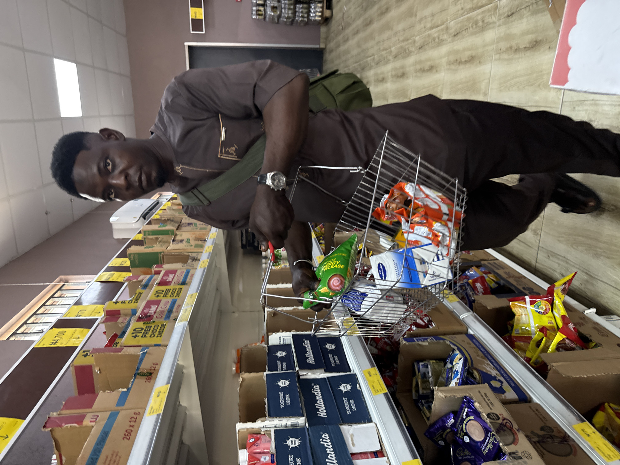 A man in a brown uniform is shopping in a supermarket. He is holding a silver wire basket filled with items, including what appears to be a green-packaged yogurt drink. The shelves in front of him contain boxes of food and drink products, like Hollandia Yogurt Drink and Cadbury Hot Chocolate. Based on the OCR, there are tags with prices and descriptions such as "+10 Free Choco Inside" visible. The location may be in Iranla, Nigeria, according to the OCR.