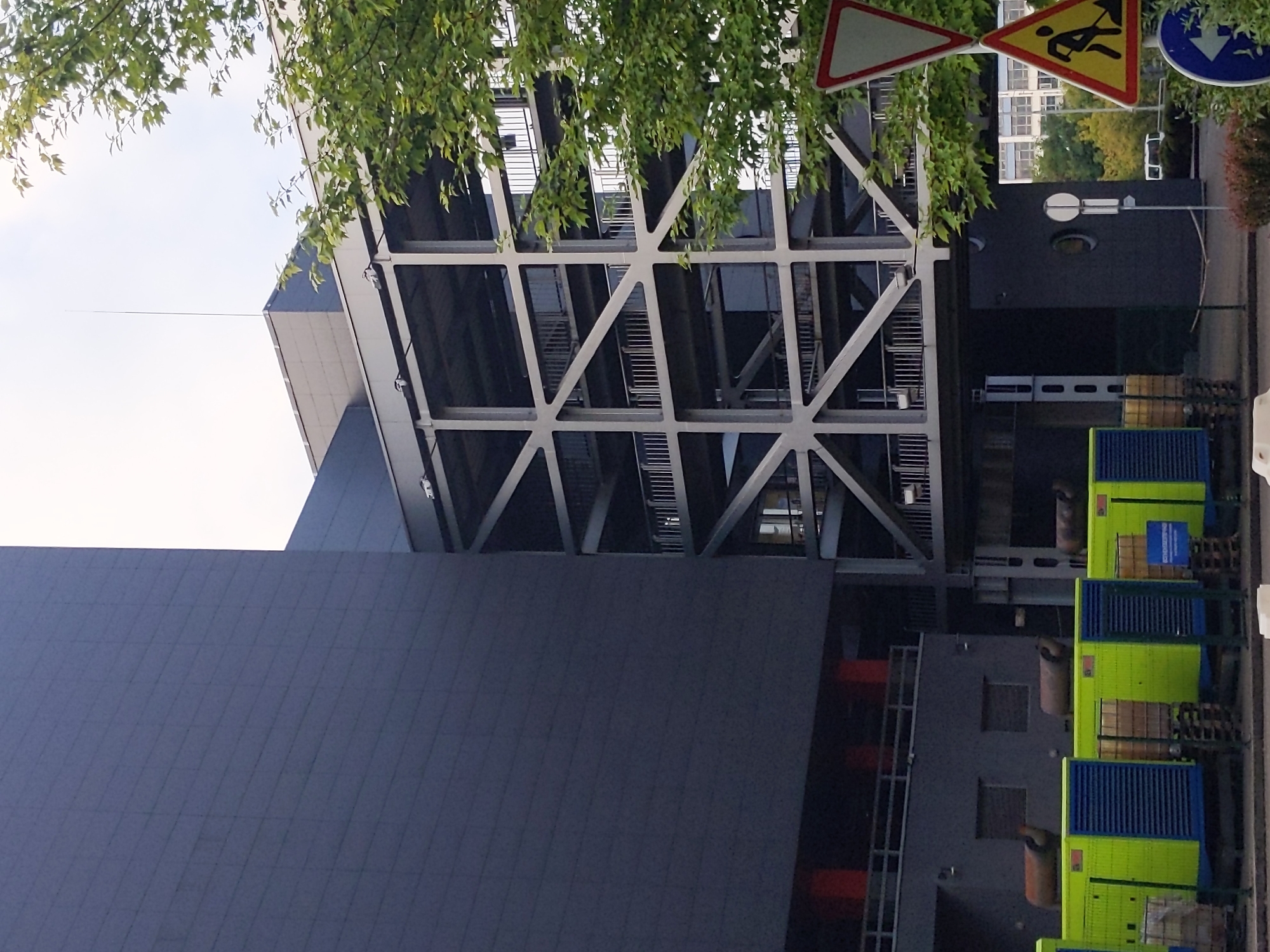 This image depicts an outdoor scene featuring a modern industrial or administrative building with a dark gray, panel-covered facade on the left. Adjacent to it, to the right, is a multi-story, open-air structure with a complex geometric metal framework, supporting walkways or platforms with railings. Green and blue generators are lined up in the foreground. In the background, there are trees, a street with a white vehicle, and a white building. Two traffic signs are visible: a triangular red and white sign and a triangular yellow and black sign with a silhouette of a person digging. The scene is set under a bright, overcast sky. The image does not provide sufficient detail to definitively place it in Kyiv, Ukraine, but the overall aesthetic is consistent with a modern urban or industrial environment. There are no people clearly visible in the image.