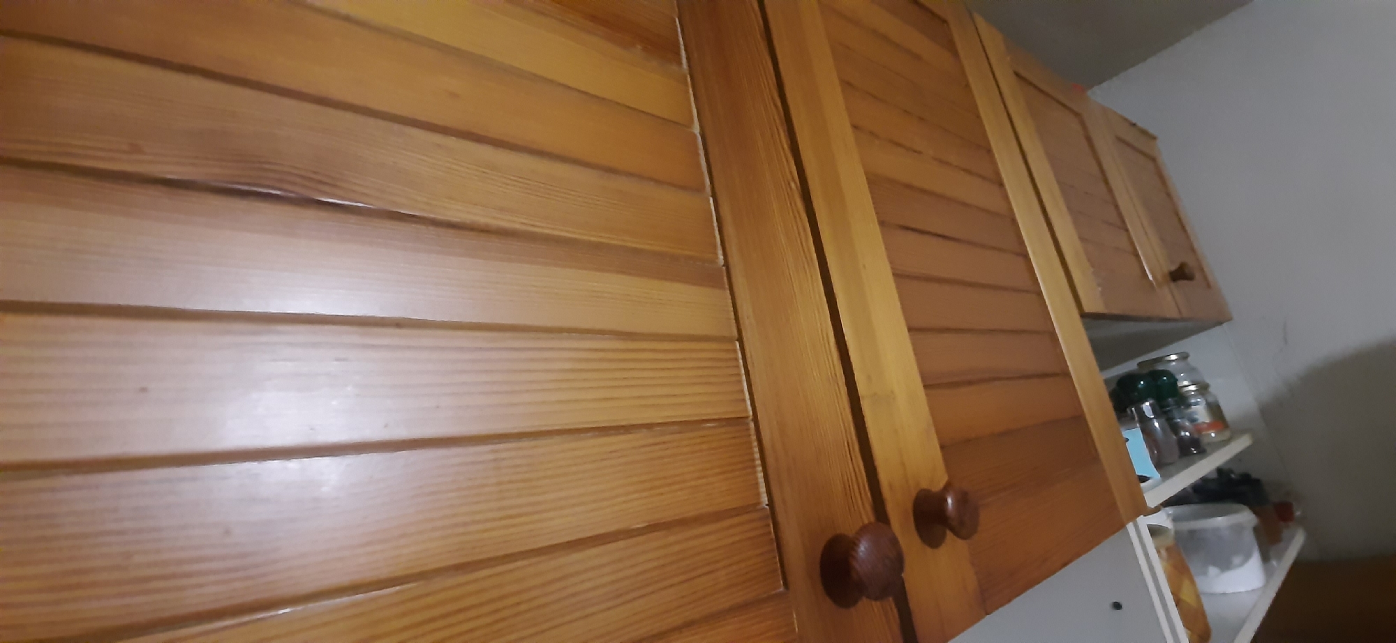 This image shows the interior of a kitchen, specifically focusing on wooden cabinetry. The cabinets are made of light-colored wood with horizontal slats. There are two cabinet doors visible on the right side of the image, each with a round wooden knob. A shelf is visible below these cabinets, holding several jars and containers, suggesting it's a food storage area. The overall environment appears to be domestic. The image's metadata does not specify a location in Yessentukskaya, Russia.