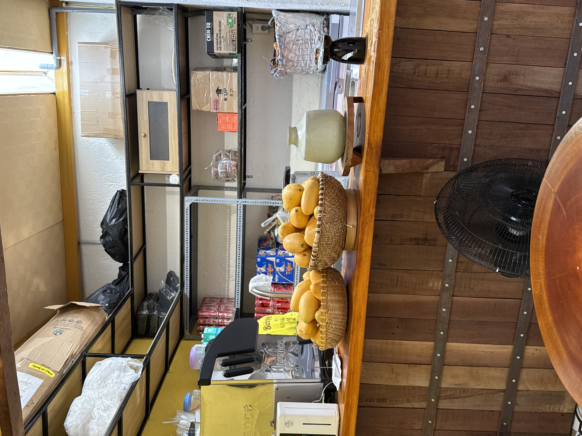 The image depicts a cluttered corner inside a building, possibly a storage or service area. Shelving units are filled with boxes, packaged goods, and various items, including a bag labeled "CHIN-SU." Near the shelves, two woven baskets filled with yellow mangoes are visible. There is a small wooden table, and an out-of-focus fan is seen nearby.
Text on the products and a sign that reads "Bánh mì Phan" indicates the location is in the Nha Trang Ward of Vietnam. A piece of equipment labeled "Elegance" and "35/3" and a white dispenser are also visible. The environment appears busy and utilitarian, suggesting a commercial setting like a restaurant or bakery.