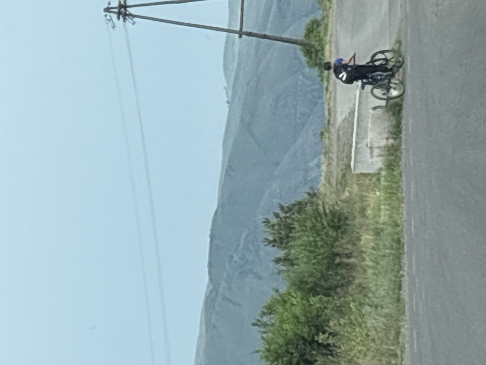 An individual wearing a backpack is on a bicycle on a paved surface with grass and hills in the background. The individual is wearing a blue shirt and dark pants. The bicycle is black with silver spokes. A power pole with wires is visible to the right of the individual. The sky is light blue and hazy. The scene appears to be a rural or undeveloped area on the outskirts of Chernogorsk, Russia.