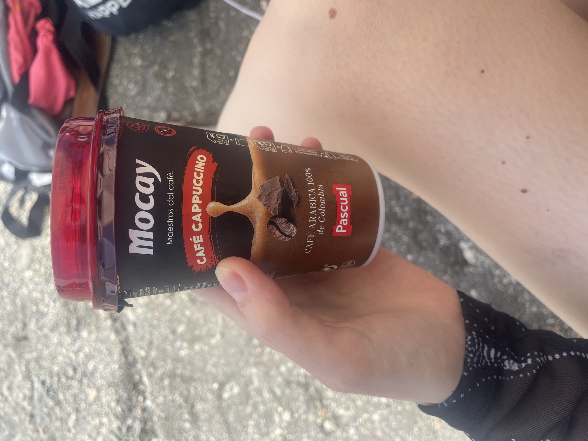 A person's hand is holding a dark brown Mocay cappuccino cup with a red lid. The cup features text that reads "Mocay Maestros del café" and "CAFÉ CAPPUCCINO CAFÉ ARABICA 100% de Colombia PASCUAL". There are images of coffee beans and chocolate chunks on the cup. The background is blurred and appears to be a pebbly surface, possibly a beach or a street. In the upper right corner, there are hints of clothing and bags, suggesting an outdoor or casual setting. The lighting is bright and natural, indicating it's daytime. The person's leg and skin are visible, with a mole on the thigh. The setting does not provide specific clues to pinpoint Almuñécar, Spain, beyond the generic description of an outdoor location.
