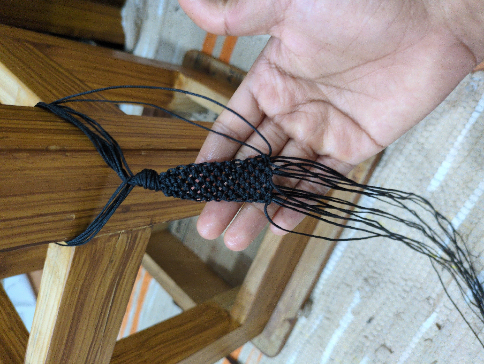A close-up, eye-level shot shows a person's hand holding a black macrame creation. The macrame is attached to a wooden structure, possibly part of a chair or stool, which is made of light-colored wood with a visible grain. The macrame itself is a black, woven design, tapering towards the bottom where multiple loose black threads hang down. The background shows a textured rug or mat with white and beige stripes. The scene is indoors, with the lighting suggesting it might be daytime. The image's focus is on the macrame and the hand, with the wooden structure and background slightly blurred. There is no discernible information about the location in Haini, India.