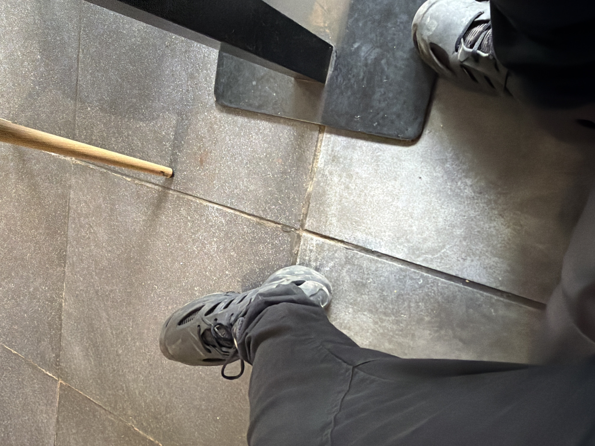 The image captures an overhead, first-person perspective of an indoor tiled floor in Sofia, Bulgaria. The primary subjects are two feet wearing dark, perforated shoes, a wooden stick, and industrial-looking fixtures.
The floor is constructed from large, square, dark gray tiles with a slightly speckled or textured surface, separated by visible grout lines. In the foreground, the photographer's left foot is prominent, clad in a dark gray or black shoe featuring multiple cut-out perforations and laces. This foot is accompanied by dark gray or black trousers. Another foot, wearing a similar dark, perforated shoe, is partially visible in the upper right portion of the frame, resting on a dark, possibly rubber, mat or base.
To the left of the foreground foot, a light brown wooden stick or dowel lies horizontally on the tiles. It is tapered at one end, which shows a slightly worn or roughened tip. This stick could be a tool, part of a game like billiards, or a simple piece of wood.
In the upper central area, a dark, rectangular structure, possibly a metal beam or part of a machine base, is visible. It casts a shadow and extends across the frame, partially obscuring the dark mat on which the second foot rests. The overall environment suggests an indoor public or commercial space, possibly a gym, workshop, or utility area.
The lighting appears artificial and consistent, providing clear visibility without indicating a specific time of day or outdoor weather. No discernible emotions are conveyed, and there is no visible text in the image.