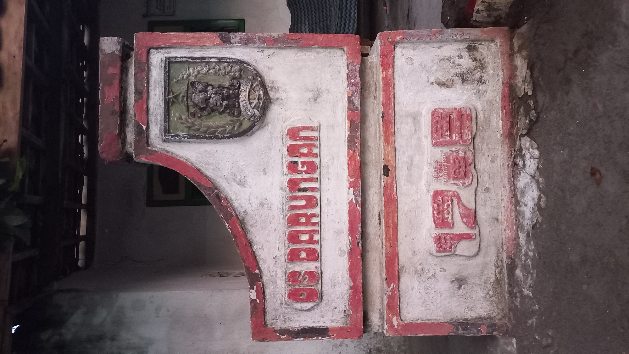 A weathered, concrete marker stands in front of a building with a rough, light-colored wall. The marker is painted white with red trim and features a coat of arms at the top. Below the coat of arms, in raised red lettering, is the text "DS BARUNGAN." The bottom section of the marker has raised red characters that appear to be a year or date, possibly "1972" or similar, though the details are obscured by wear. The ground in front of the marker is rough, gray concrete. The background shows a dark, covered area, possibly a roof or overhang, with wooden beams. To the right, a glimpse of a green-framed window and a dark, textured object, possibly furniture, is visible. The overall impression is of a historical or administrative marker in a somewhat neglected outdoor setting in Pare, Indonesia.