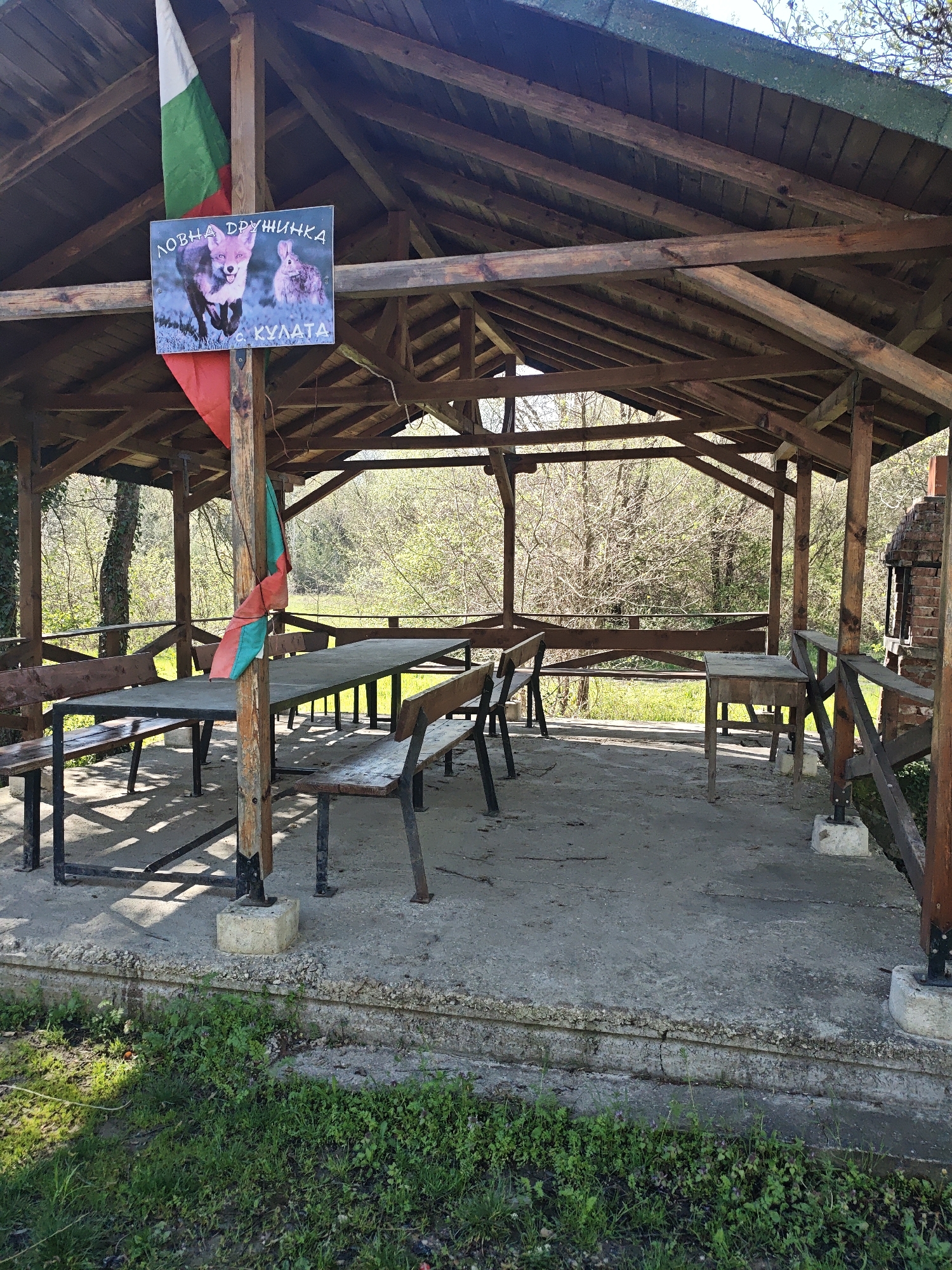 The image depicts an outdoor covered picnic area in an unidentified location. The structure is a wooden gazebo with a table and benches inside. A partially torn Bulgarian flag is attached to one of the support posts. A sign with an image of a fox and rabbit and the text "ЛОВНА ДРУЖИНКА с КУЛАТА" is also affixed to the structure. The surrounding area appears to be wooded, with visible trees and vegetation.