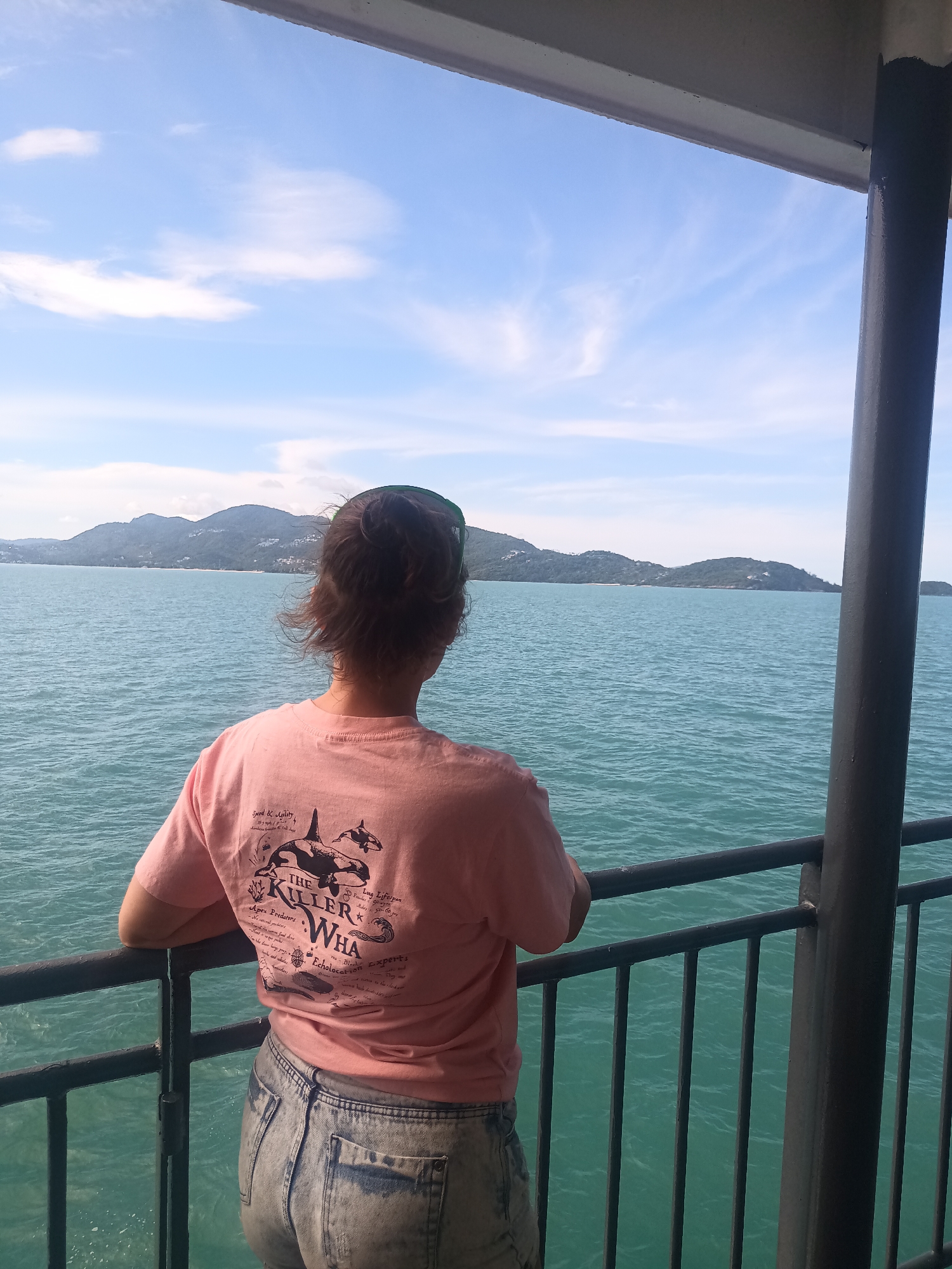 A female figure is standing with her back to the camera, gazing out over the water from what appears to be a boat or ferry. She is wearing a light pink t-shirt with a graphic print of killer whales and faded denim shorts. Her hair is gathered up, and she has a pair of sunglasses on top of her head.
The woman is leaning slightly against a dark metal railing, which has vertical bars forming a safety barrier. Behind her is a body of water with visible ripples, stretching out to an island in the distance. The sky above is blue with scattered white clouds.
The scene is partially sheltered by a structure with a dark gray column to the right of the frame. The location is Baan Thong Po, Thailand.