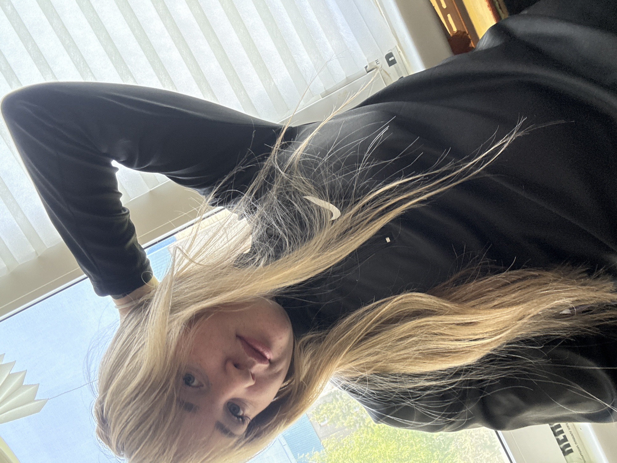 A young woman with blonde hair wearing a black long-sleeved Nike shirt is positioned upside down in the frame. She appears to be indoors, with a window featuring white blinds visible in the background. Outside the window, green trees and a building can be seen. The location in the city of Марковский, Russia, cannot be confirmed from the image.