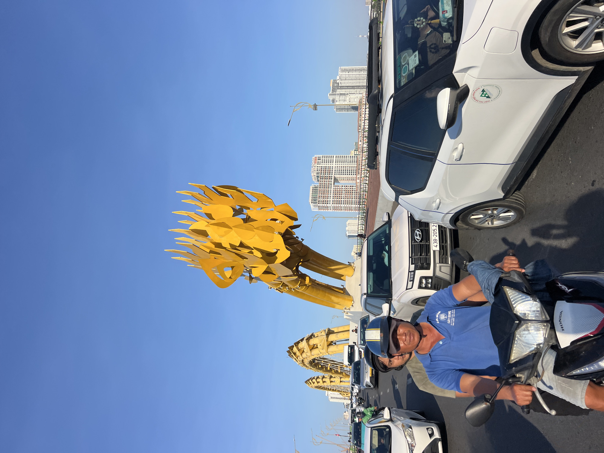 The image depicts a street scene on a bridge in Da Nang, Vietnam, featuring the Dragon Bridge. The Dragon Bridge is a prominent landmark, characterized by its striking yellow, dragon-shaped sculpture. In the foreground, two individuals are riding a motorcycle, with the driver wearing a blue polo shirt and a helmet. They are part of a traffic jam, with several cars and other motorcycles around them. The vehicles are moving along the bridge, under a clear blue sky. In the background, several tall buildings and a street lamp are visible, indicating an urban environment. The overall scene is that of a bustling city, with traffic and iconic architecture.