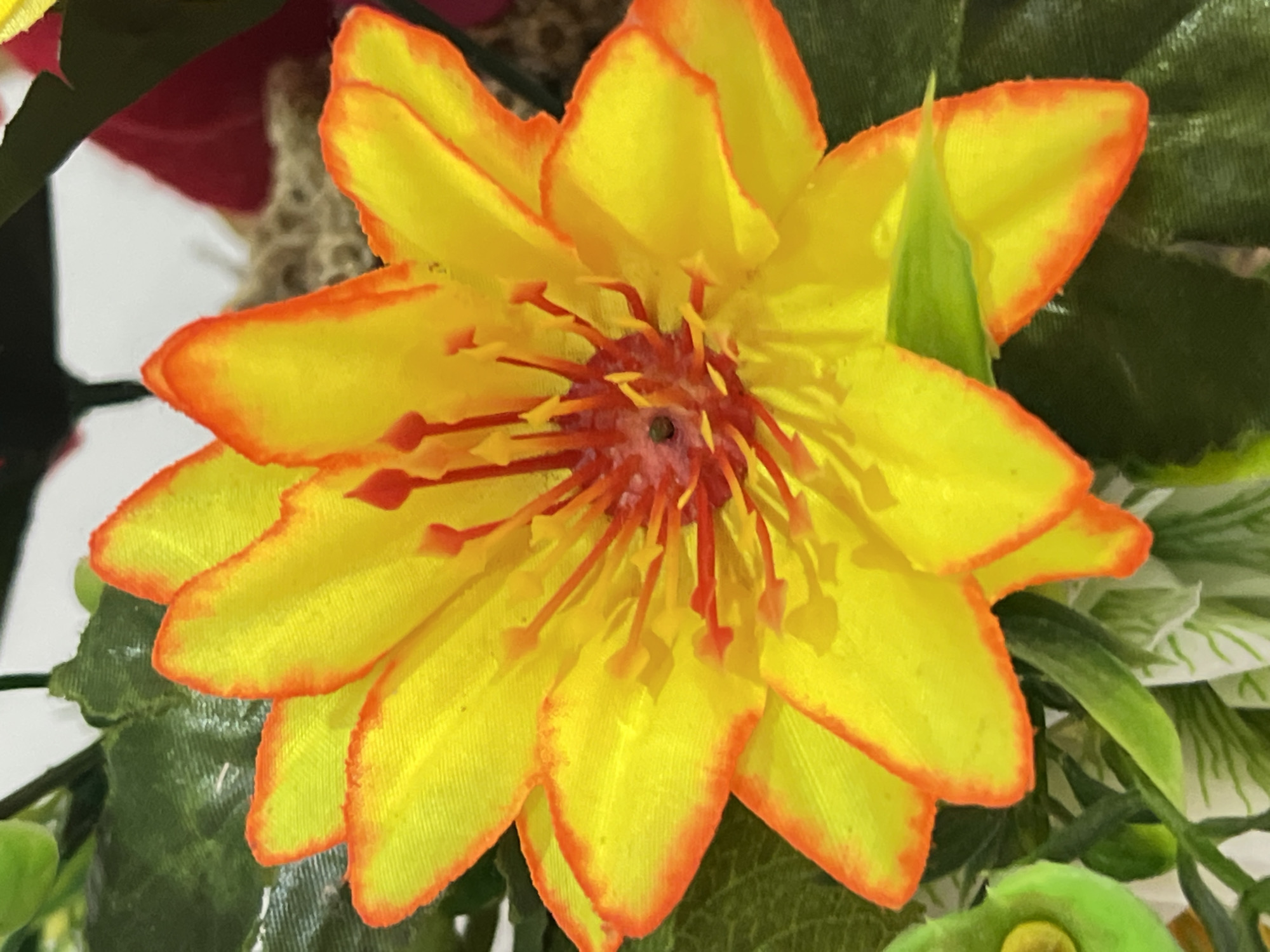 A close-up, eye-level, vertical shot captures a vibrant yellow artificial flower with orange-tinged petal edges. The flower's center features red stamen-like structures, and the petals appear slightly layered. Surrounding the flower are dark green artificial leaves and hints of other floral elements in red and brown in the blurred background. The image is taken indoors, potentially in a home or a commercial setting, in Denpasar, Indonesia, possibly as part of a decorative arrangement. There are no people or animals present, and no discernible actions are taking place beyond the static display of artificial flora. The lighting appears to be artificial, casting soft shadows on the petals.