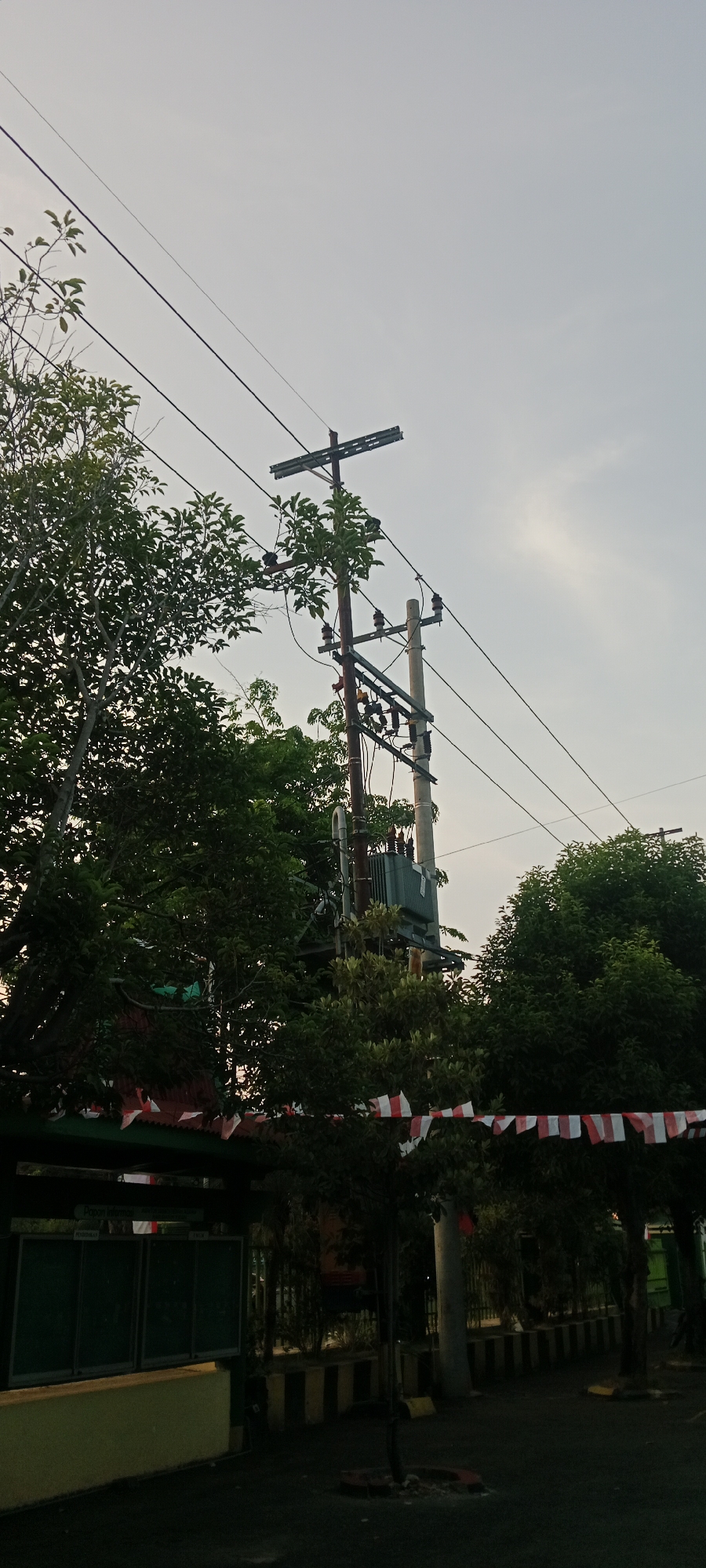 The image shows a utility pole with electrical wires and a transformer, surrounded by lush green trees. Red and white pennant banners are strung across the scene, suggesting a festive or patriotic occasion. In the foreground, a dark paved area is visible, with some yellow and black striped barriers along the edge of a building. The sky is overcast, indicating a cloudy day. The scene appears to be in an outdoor urban or suburban setting. There are no people visible in the image.