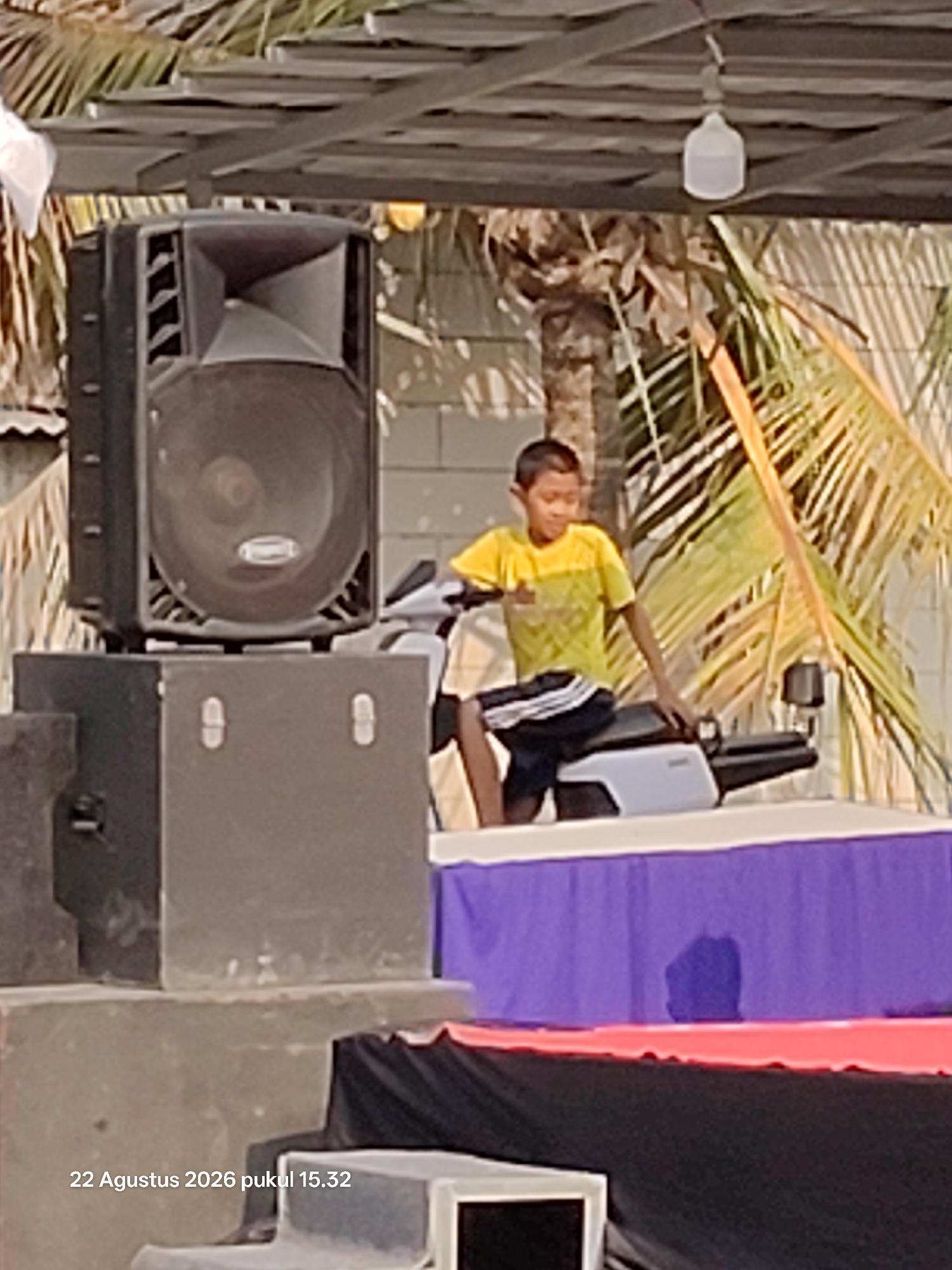 An outdoor scene in Wedi, Indonesia, features a young boy in a yellow and green t-shirt and black shorts sitting on a white motorcycle. He is positioned on a stage or platform covered with a purple cloth. To the left of the boy, a large black speaker stands on a grey pedestal. The background shows a palm tree and a wooden structure overhead, with a lightbulb hanging from it. The date and time stamp "22 Agustus 2026 pukul 15.32" is visible in the lower left corner.