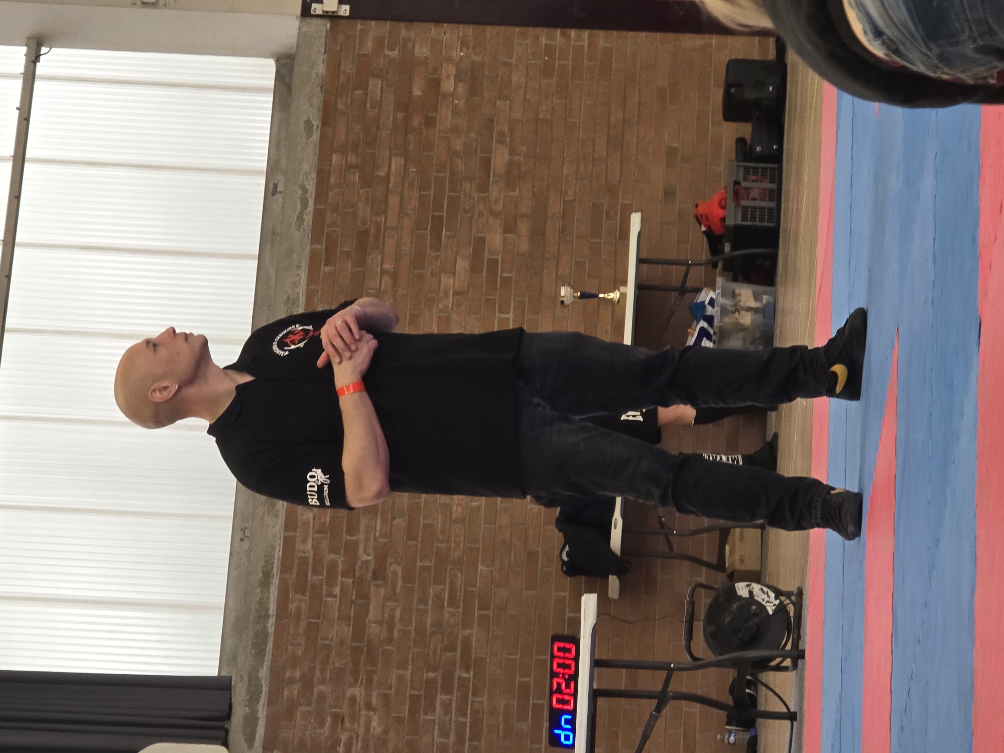 The image depicts an indoor setting, possibly a gymnasium or martial arts training center in Sainghin-en-Weppes, France. A bald man wearing a black polo shirt with visible logos on the left sleeve and chest, dark jeans, and black sneakers with yellow swooshes stands with his arms crossed. He is positioned in front of a brick wall and a white wall. A timer showing "00:20 up" in blue and red digital display is visible. In the background, there is a blue and red striped mat, gym equipment, and what appears to be a trophy on a table.