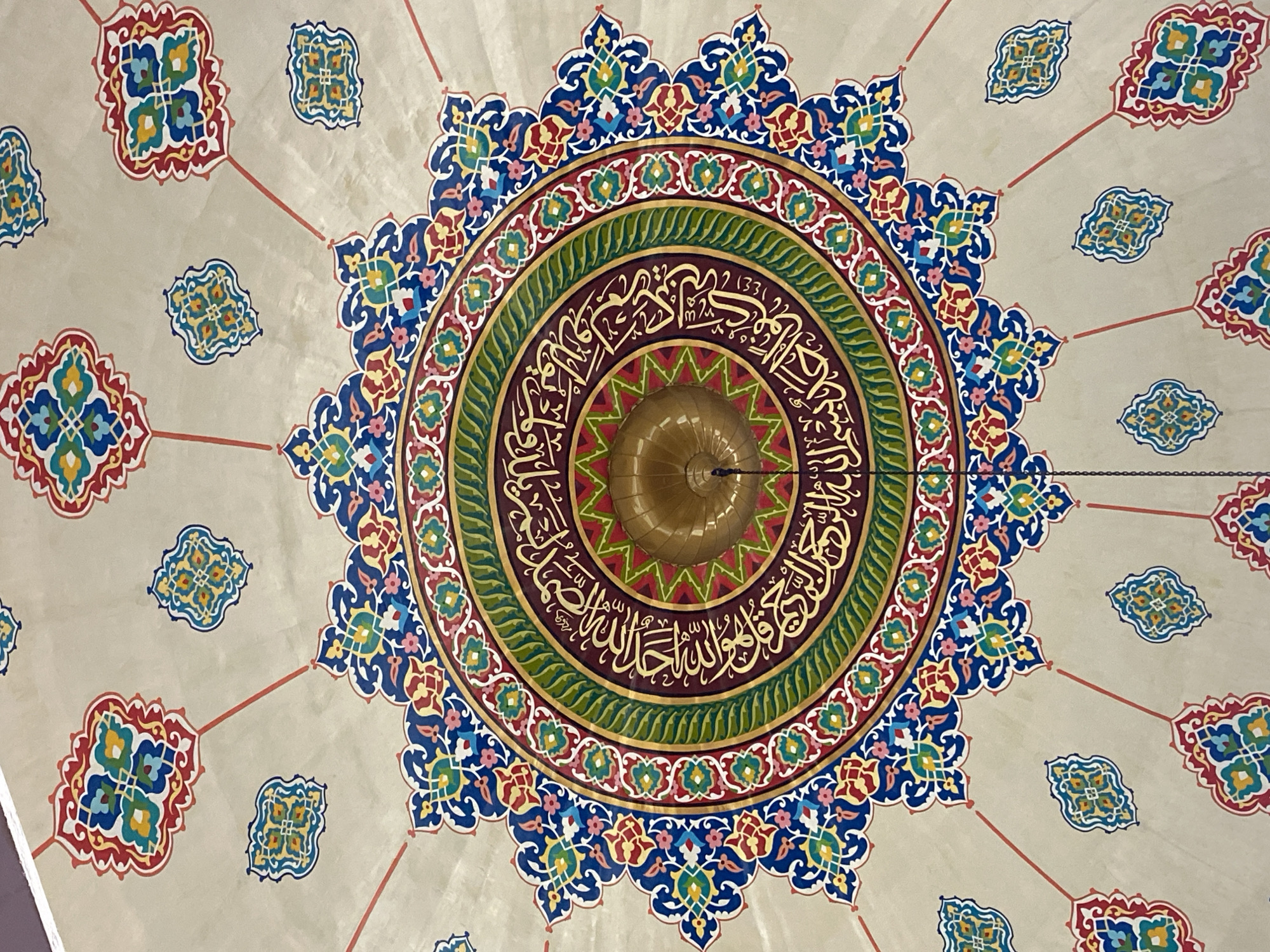 The image shows a detailed view of the ornate ceiling of a mosque. The ceiling features intricate geometric patterns and Arabic calligraphy in vibrant colors of blue, red, green, yellow, and gold against a neutral beige background. In the center of the dome, a golden fixture hangs, likely a light source or part of the mosque's architectural design. Surrounding the central fixture are concentric rings of colorful decorative elements and golden Arabic script. Radiating from the center, thin red lines extend outwards, connecting to smaller decorative motifs on the ceiling. These motifs are also composed of geometric shapes and rich colors, creating a sense of symmetry and grandeur. The overall impression is one of meticulous craftsmanship and religious artistry. This ceiling is located in the Masjid Agung Islamic Center Lhokseumawe, in the city of Lhokseumawe, Indonesia. There are no people or animals visible in this image.