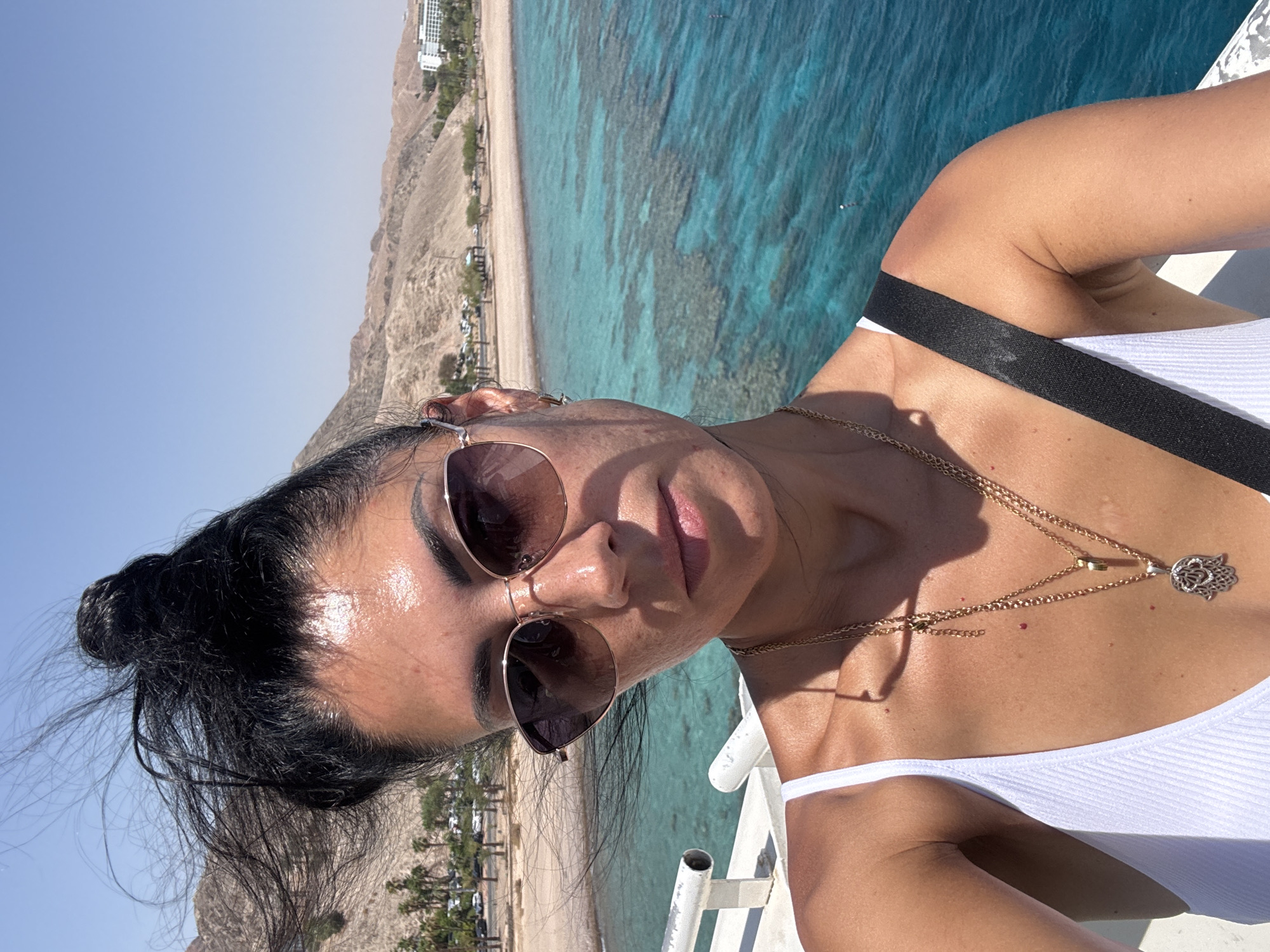 The image is a selfie taken by a woman in Eilat, Israel, with the Red Sea in the background. The woman has dark hair styled in a bun, with strands blowing in the wind. She is wearing sunglasses, a white tank top, and two gold necklaces. The background shows a calm turquoise sea with visible coral reefs, a sandy beach with palm trees, and arid, rocky hills. Buildings, possibly hotels, are visible on the coastline in the distance. The scene suggests a sunny day in a coastal resort area.